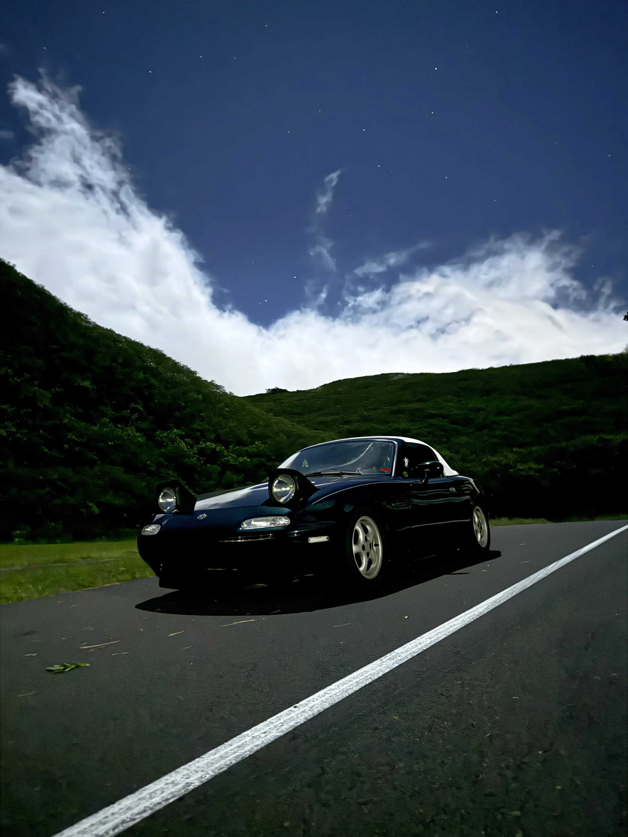A classic convertible sports car with pop-up headlights parked on a road, surrounded by green hills and a partly cloudy night sky with stars.