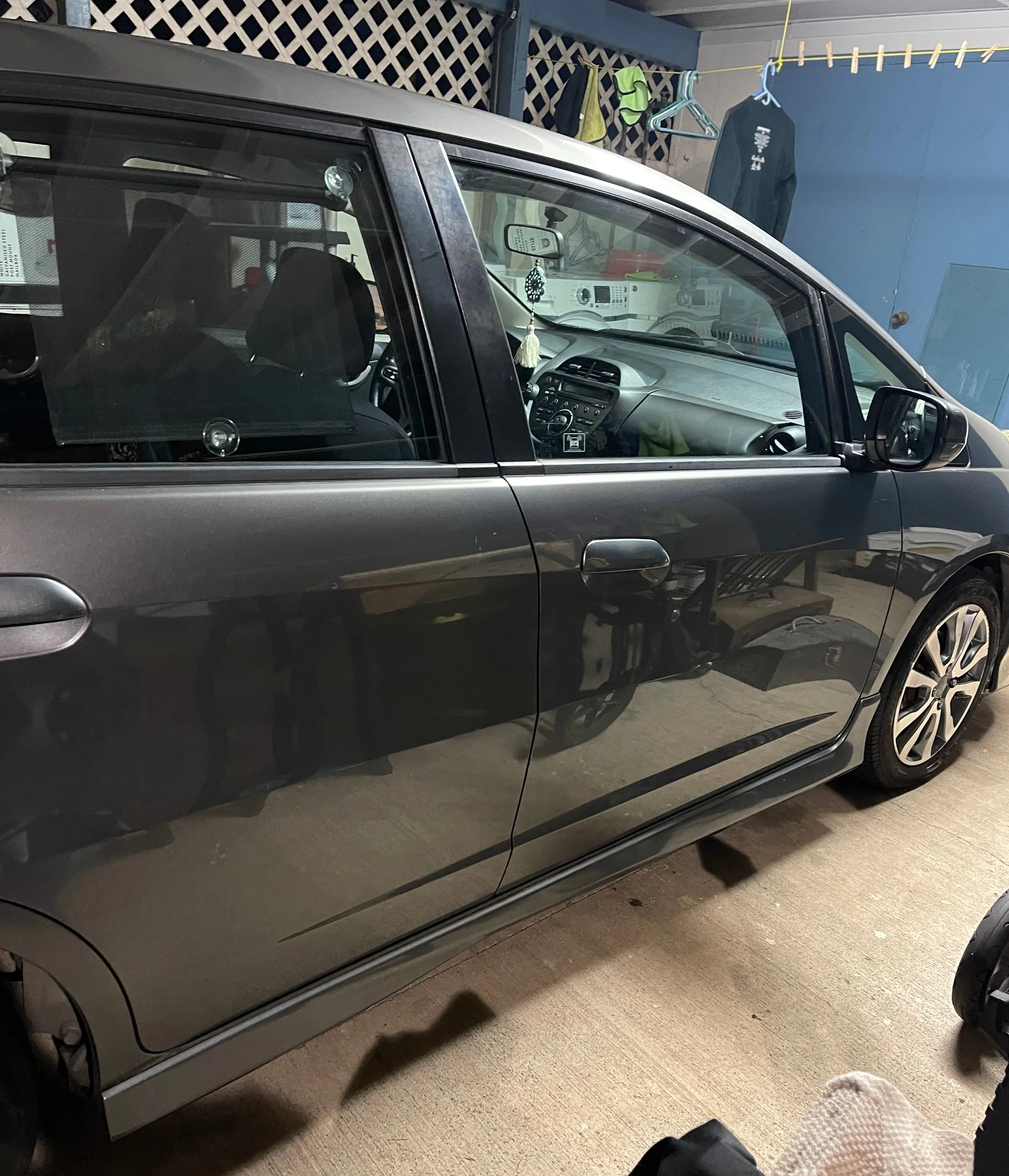 Gray car parked in a garage next to hanging clothes and a stroller.