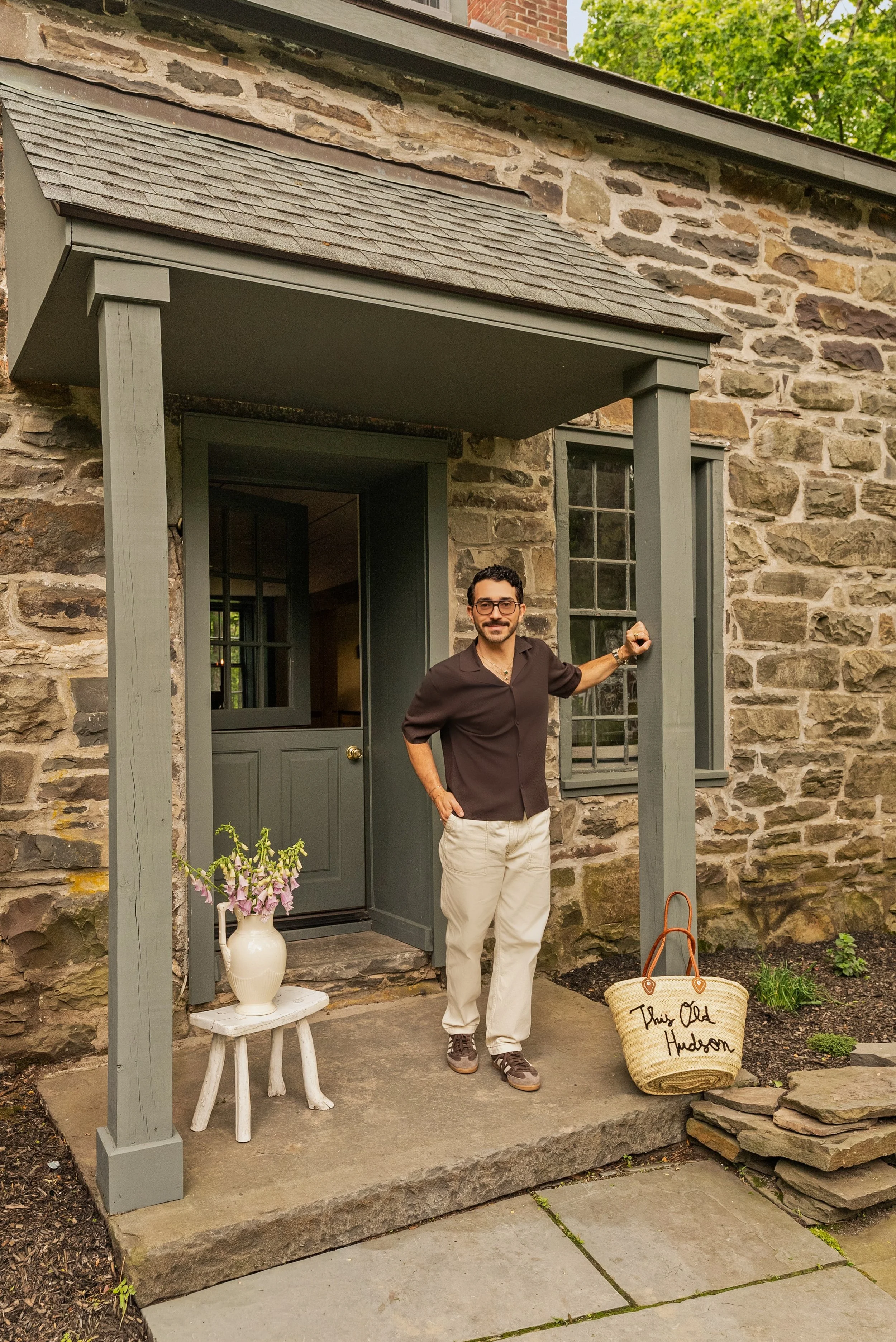 A man dressed in a brown shirt and cream pants stands on a small porch in front of a stone house. He is smiling and holding the doorframe. There is a white vase with pink flowers on a white stool to his left and a large straw bag with the words "This Old Hudson" on it placed on the ground to his right. The porch has gray painted wooden supports and a small overhang. A lush green tree is visible in the background.