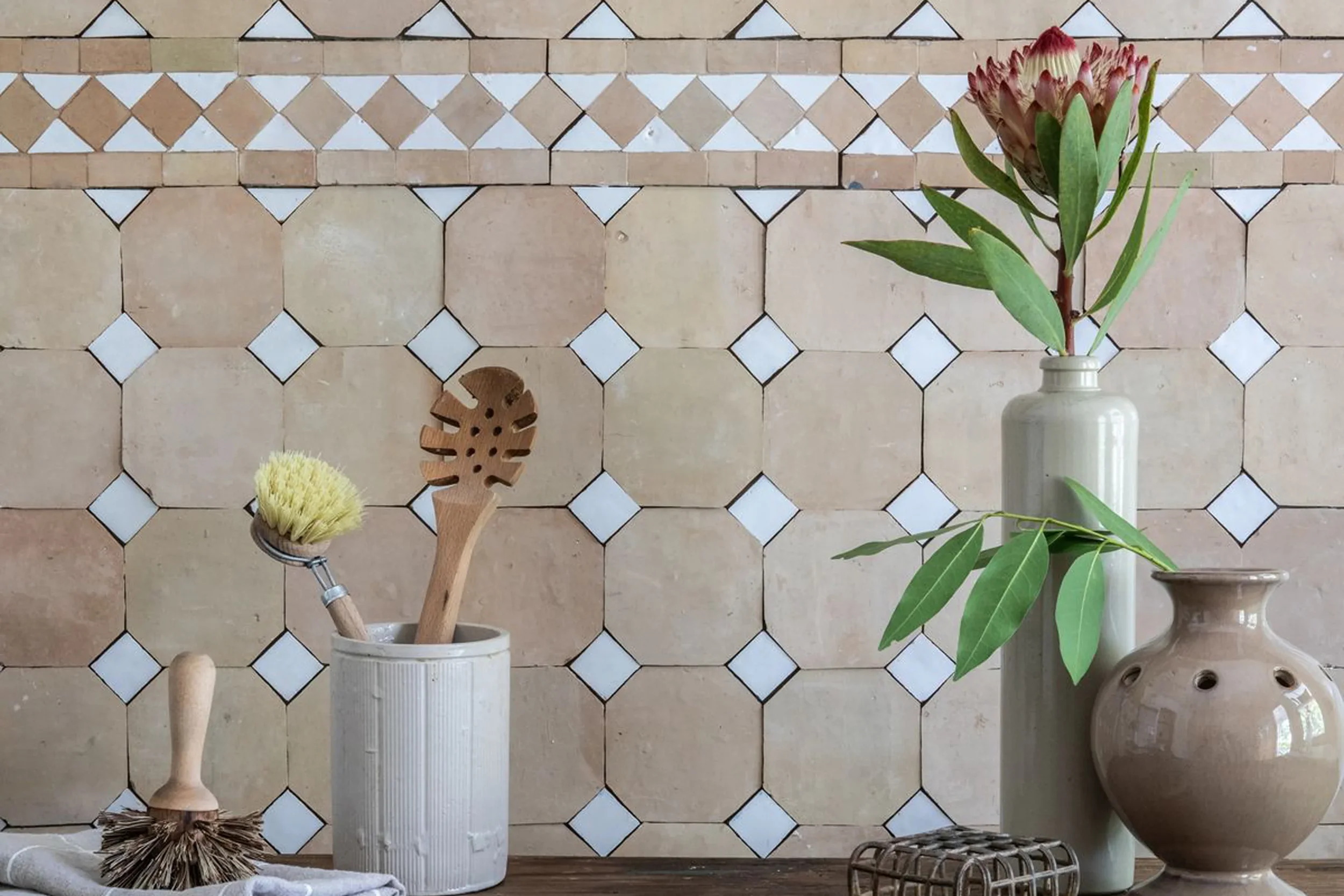 Decorative bathroom wall with beige tile pattern, a tall white vase with green leaves, a smaller beige vase, a wooden dishbrush, and a natural bristle scrub brush on a wooden surface.