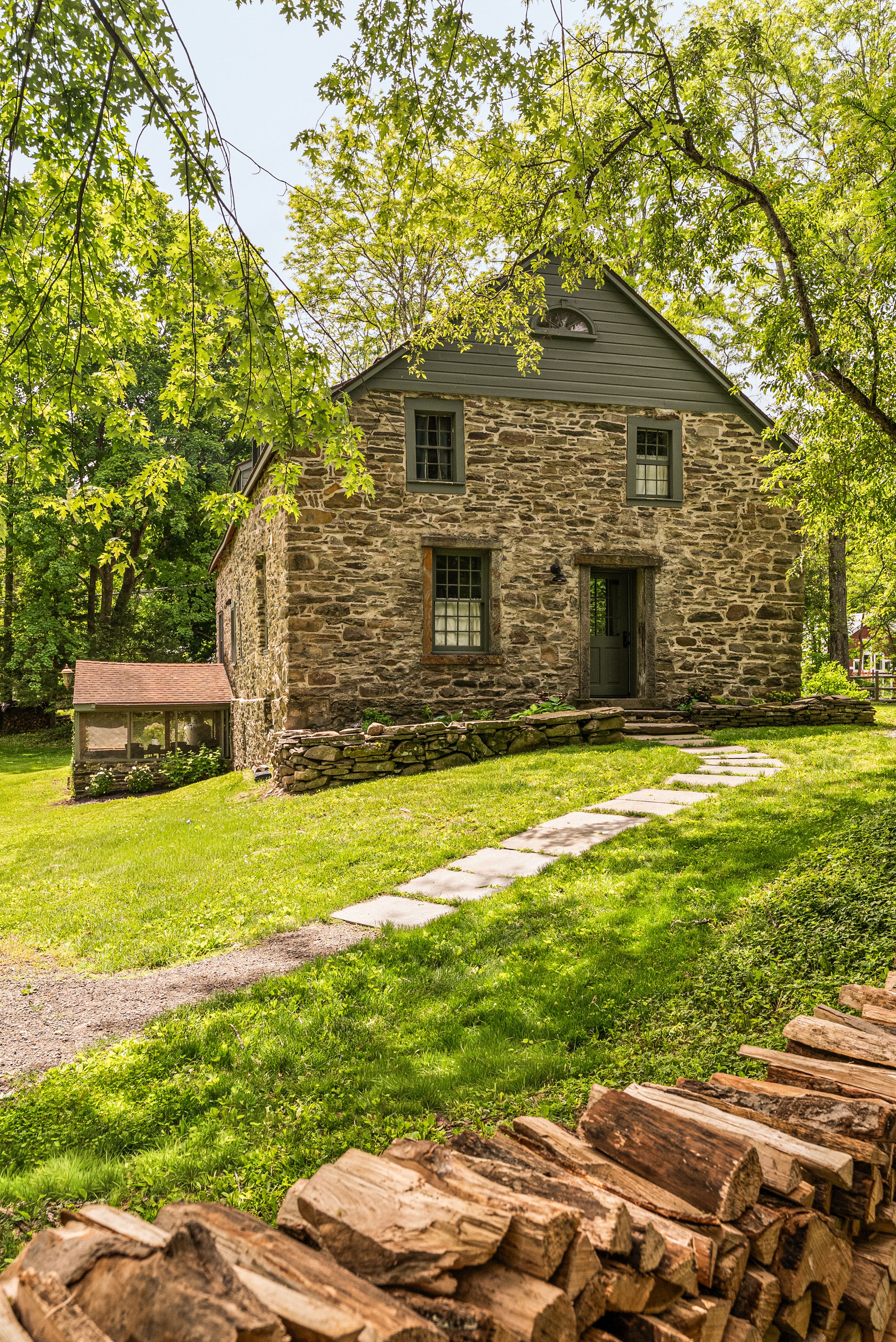 A stone house surrounded by green trees with a grassy front yard, a stone pathway leading to the door, and a pile of firewood in the foreground.