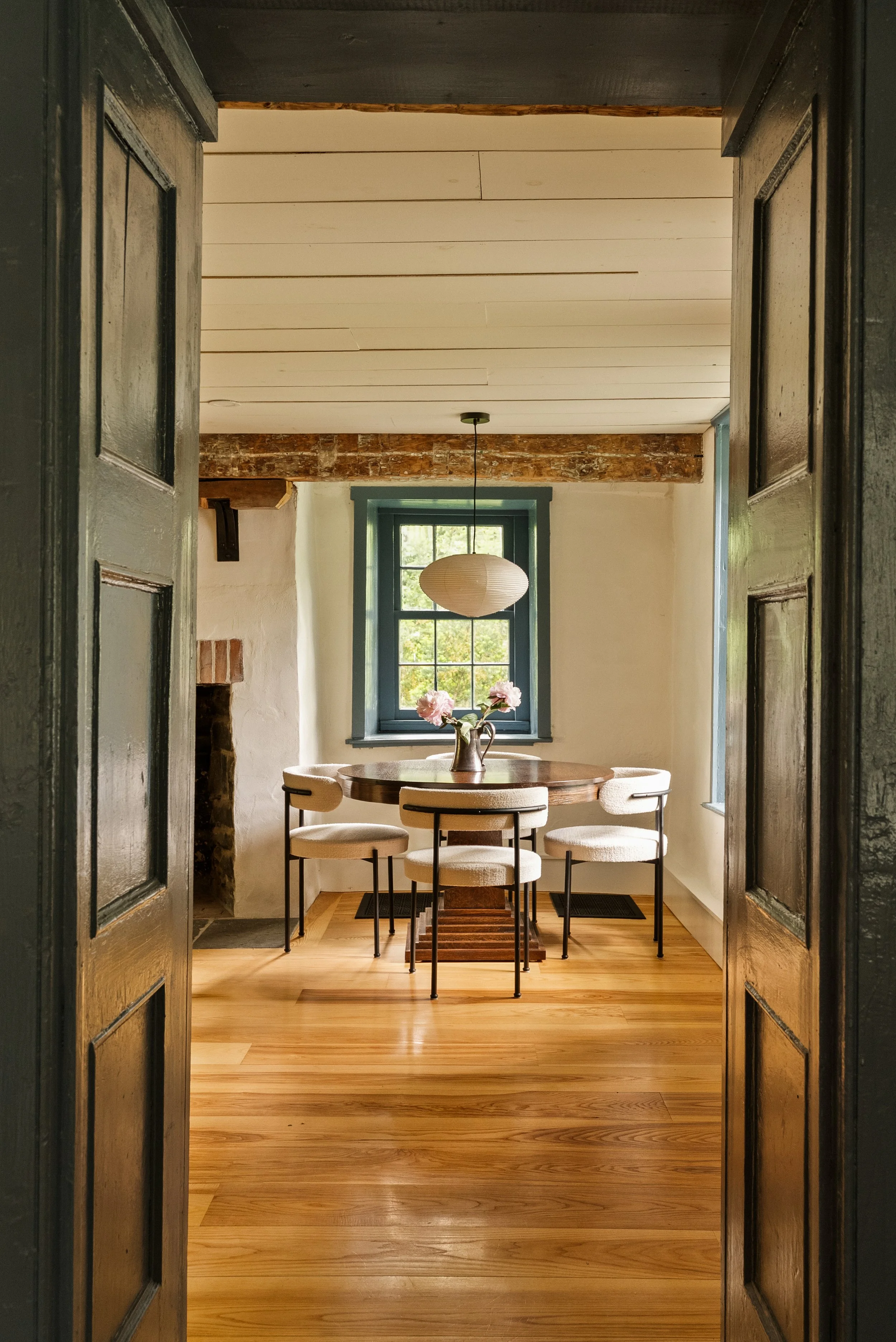 View through open black wooden doors into a dining room with a round wooden table, six beige upholstered chairs, a brick fireplace, a window with green trim, and a hanging paper lantern light fixture.