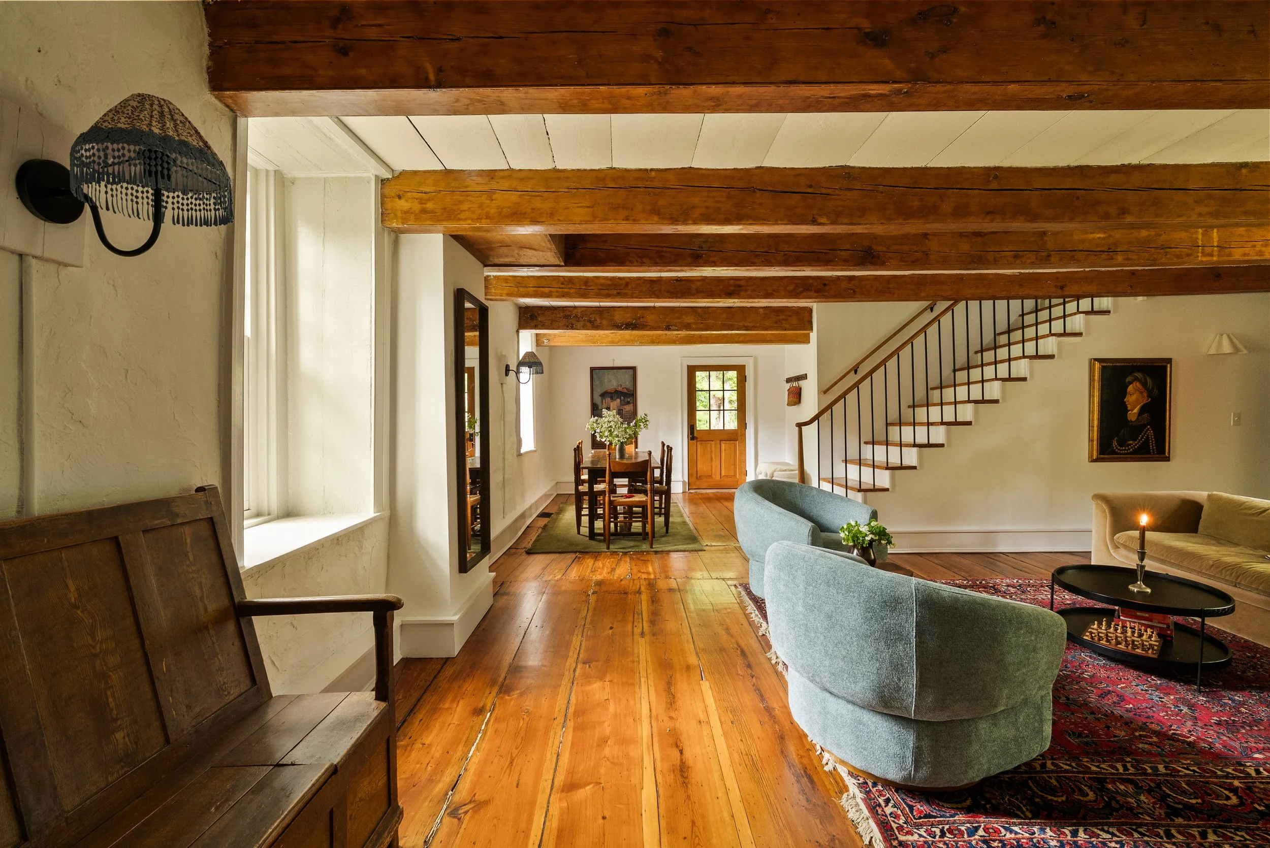 Living room with wooden floors, exposed ceiling beams, a staircase with black railing, a dining table with chairs, a sofa, armchairs, a lamp, and framed artwork on the walls.