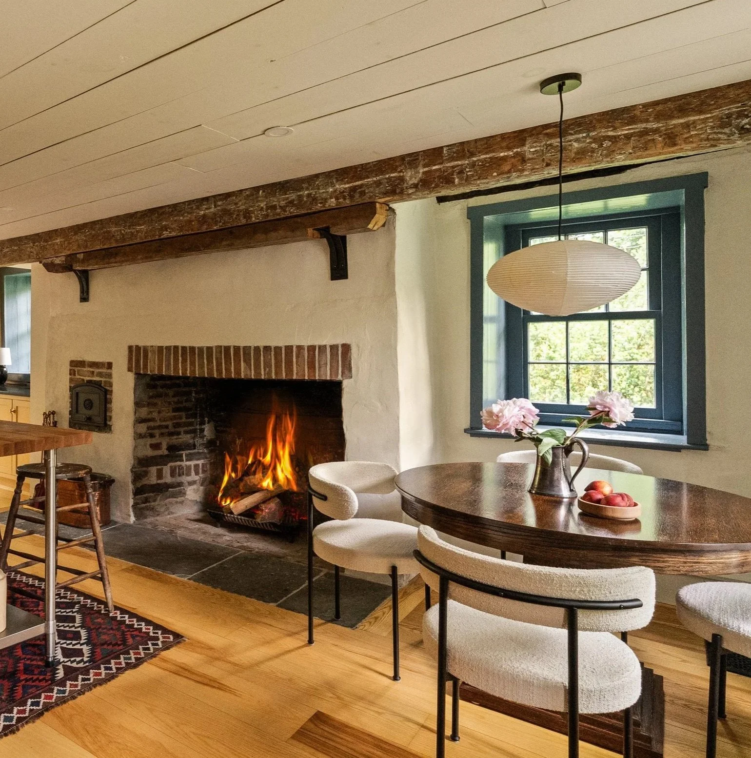 A cozy living room with a fireplace, a round wooden dining table with four cream upholstered chairs, a vase with pink flowers, a bowl of apples, and a window showing greenery outside. A paper lantern light hangs from the ceiling.