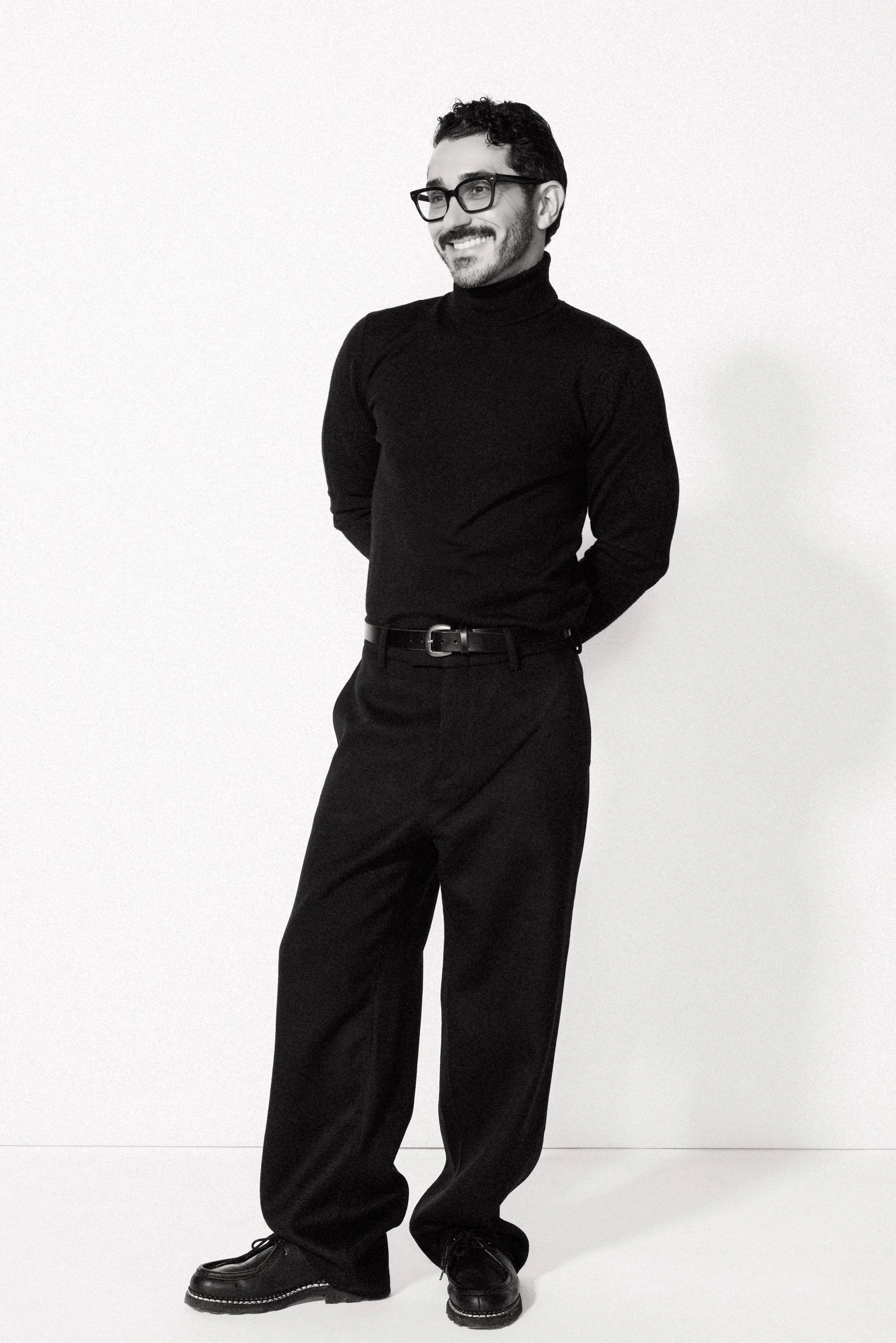 A man with dark hair and glasses wearing a black turtleneck sweater, wide-leg black trousers, and black shoes, standing against a plain white background smiling with his hands clasped behind his back.