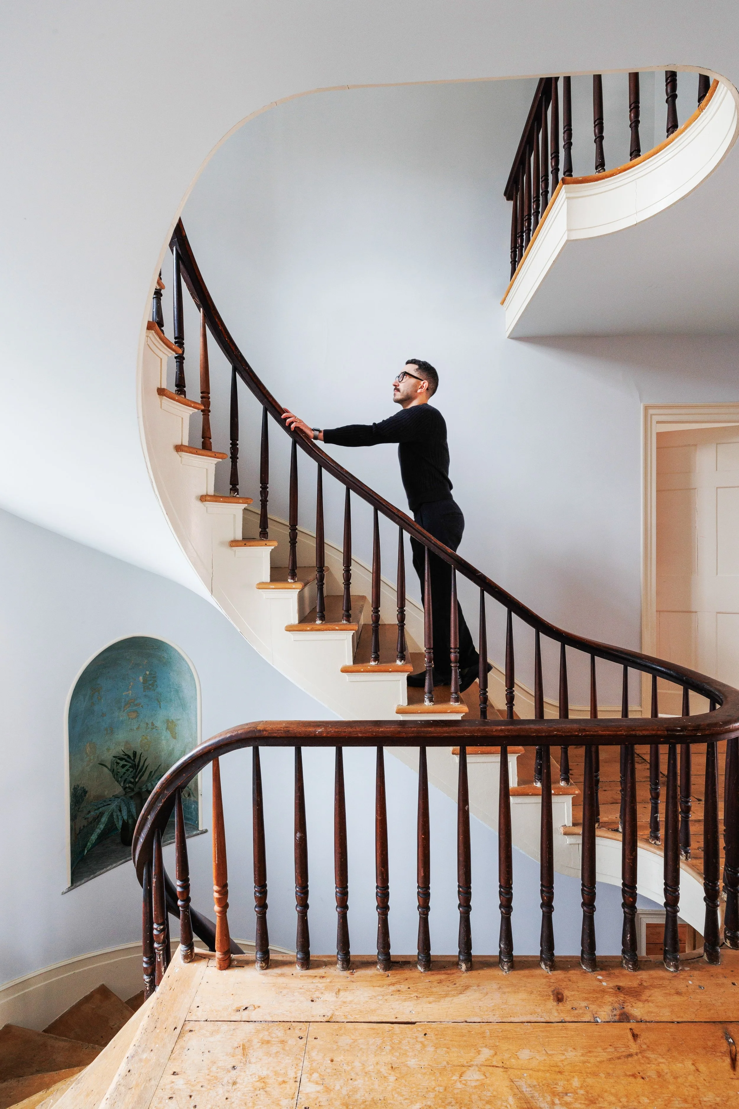 A man standing on a wooden staircase with dark wooden railings, reaching out with one hand. The staircase curves upward to the right and is inside a house with light-colored walls and a small decorative niche with plants underneath. The man is wearing a black sweater and glasses.