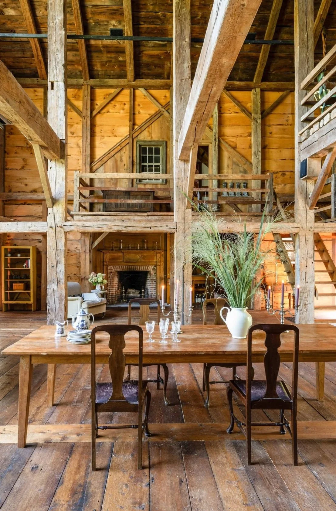 Rustic wooden interior of a house with a high ceiling, large fireplace, wooden dining table set with glasses, plates, and a large white vase with tall grasses, and a loft above with railings and stairs.
