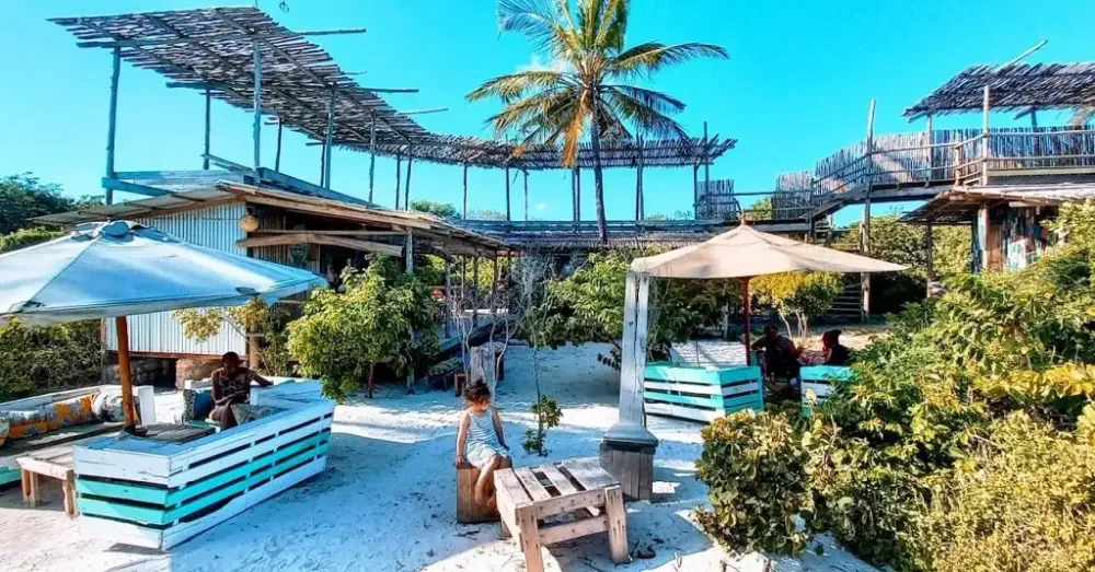 Beachside outdoor seating with umbrellas, a central palm tree, sandy ground, and a mix of wooden and bamboo structures with lush greenery.