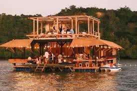 People enjoying a multi-level floating house on a river with a natural wooded background.