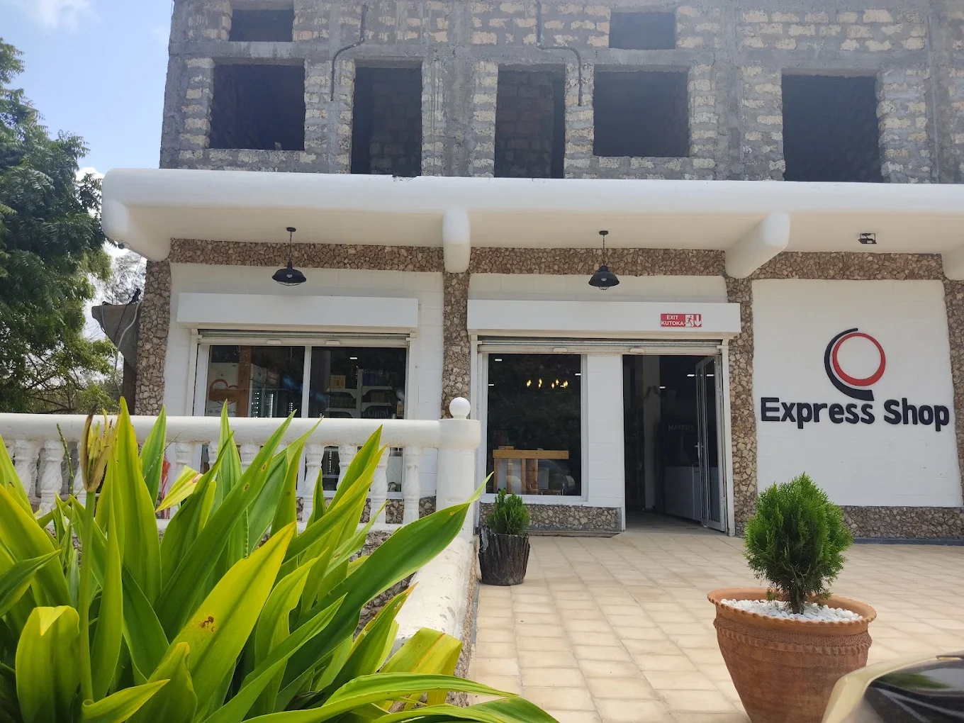 Exterior view of a small shop named 'Express Shop' with large windows, potted plants, and greenery surrounding the entrance. The building has a stone and white exterior with a sign displaying the shop's name and logo.