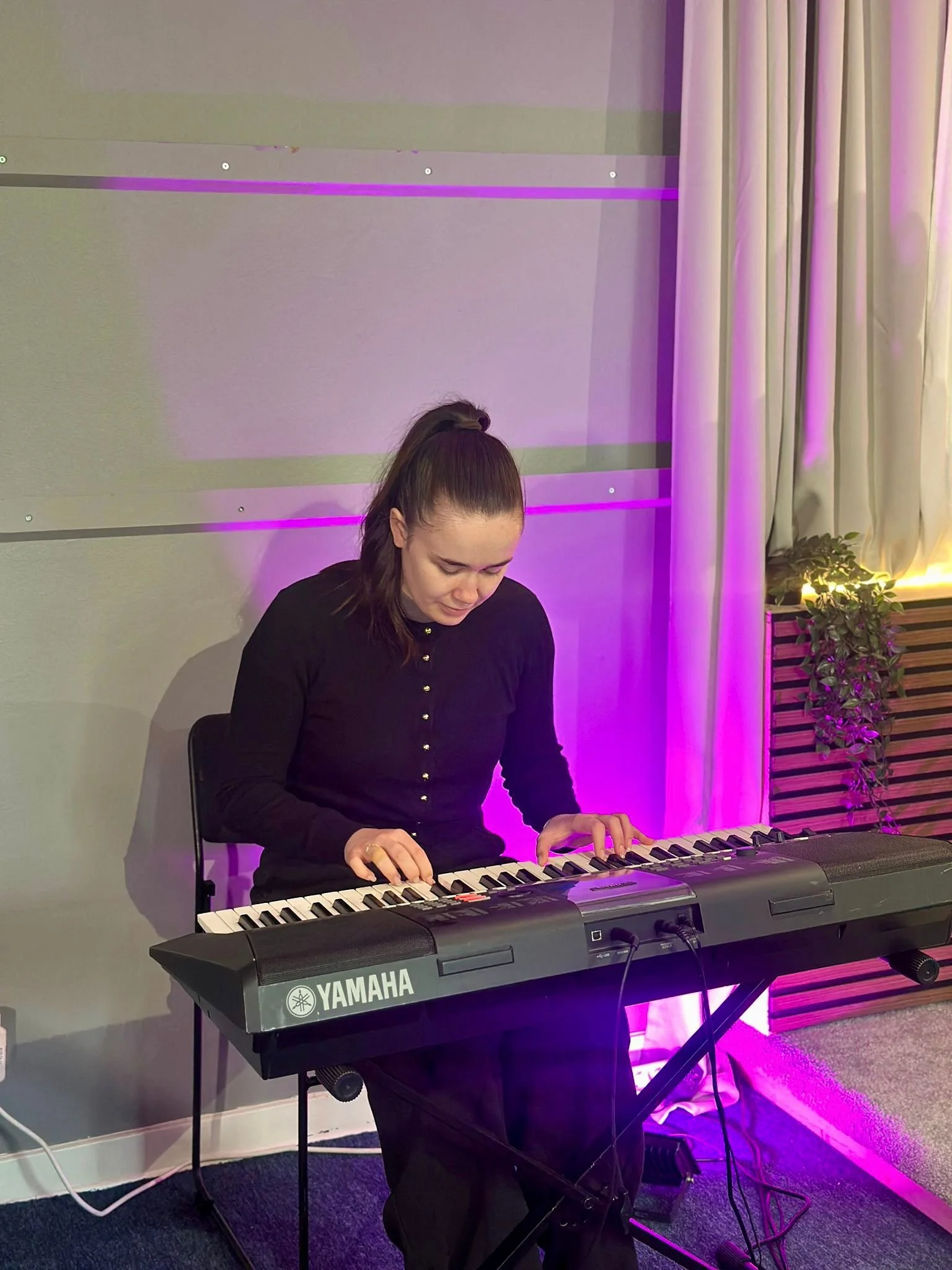 A woman with long brown hair, wearing a black shirt, playing a Yamaha electronic keyboard in a softly lit room with purple and pink lighting and cream-colored curtains.