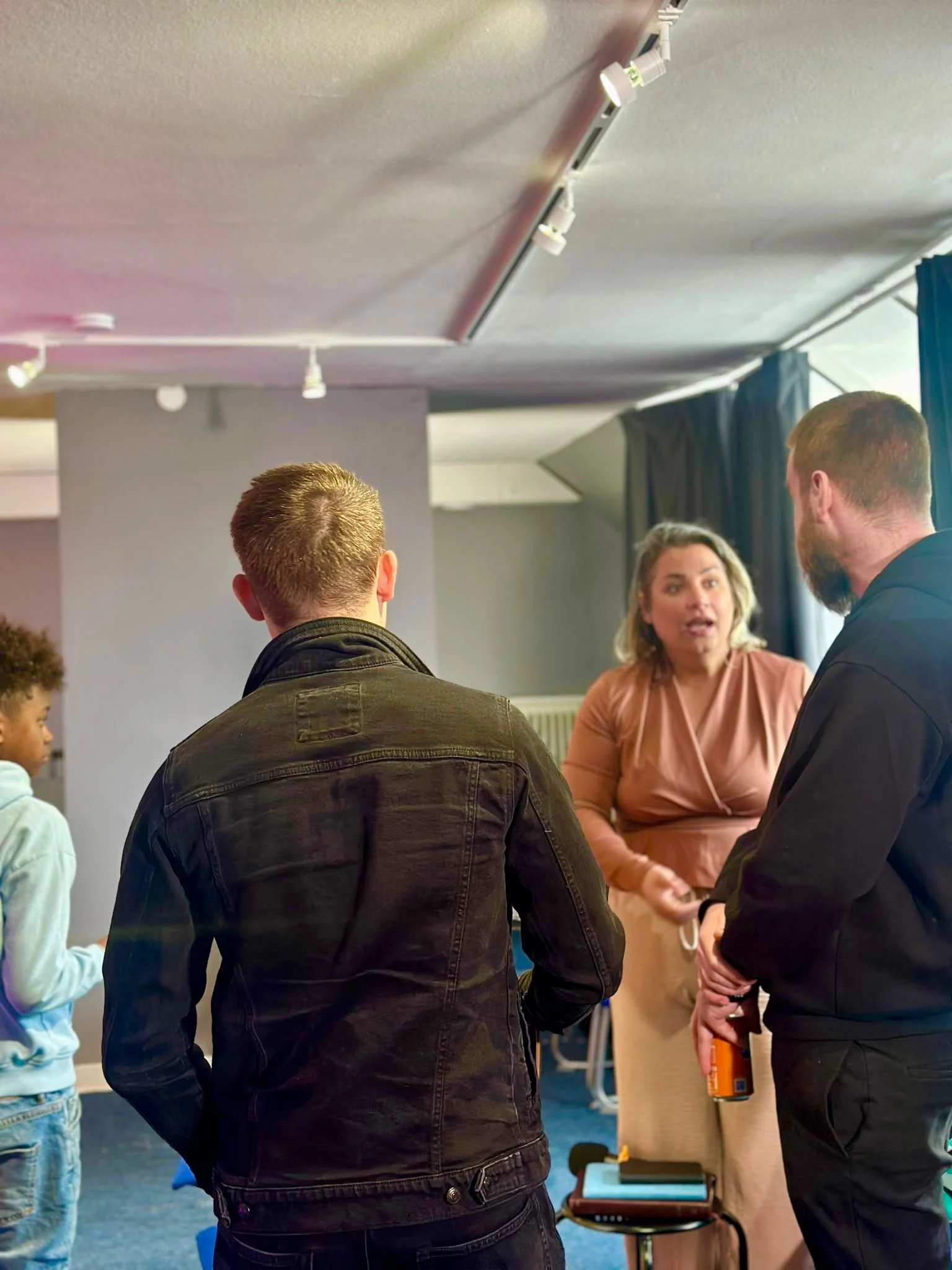 A group of four people engaged in conversation in an indoor setting, with the woman in a pink dress talking and gesturing with her hands.