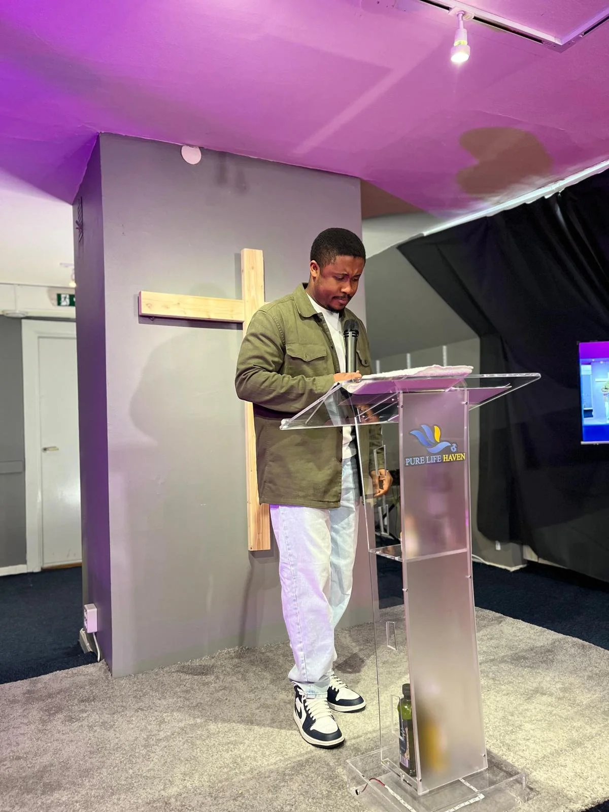 A man standing behind a clear podium with a Bible, speaking into a microphone at a church service. The podium has a logo and the words "Pure Life Haven." A large wooden cross is mounted on the wall behind him.