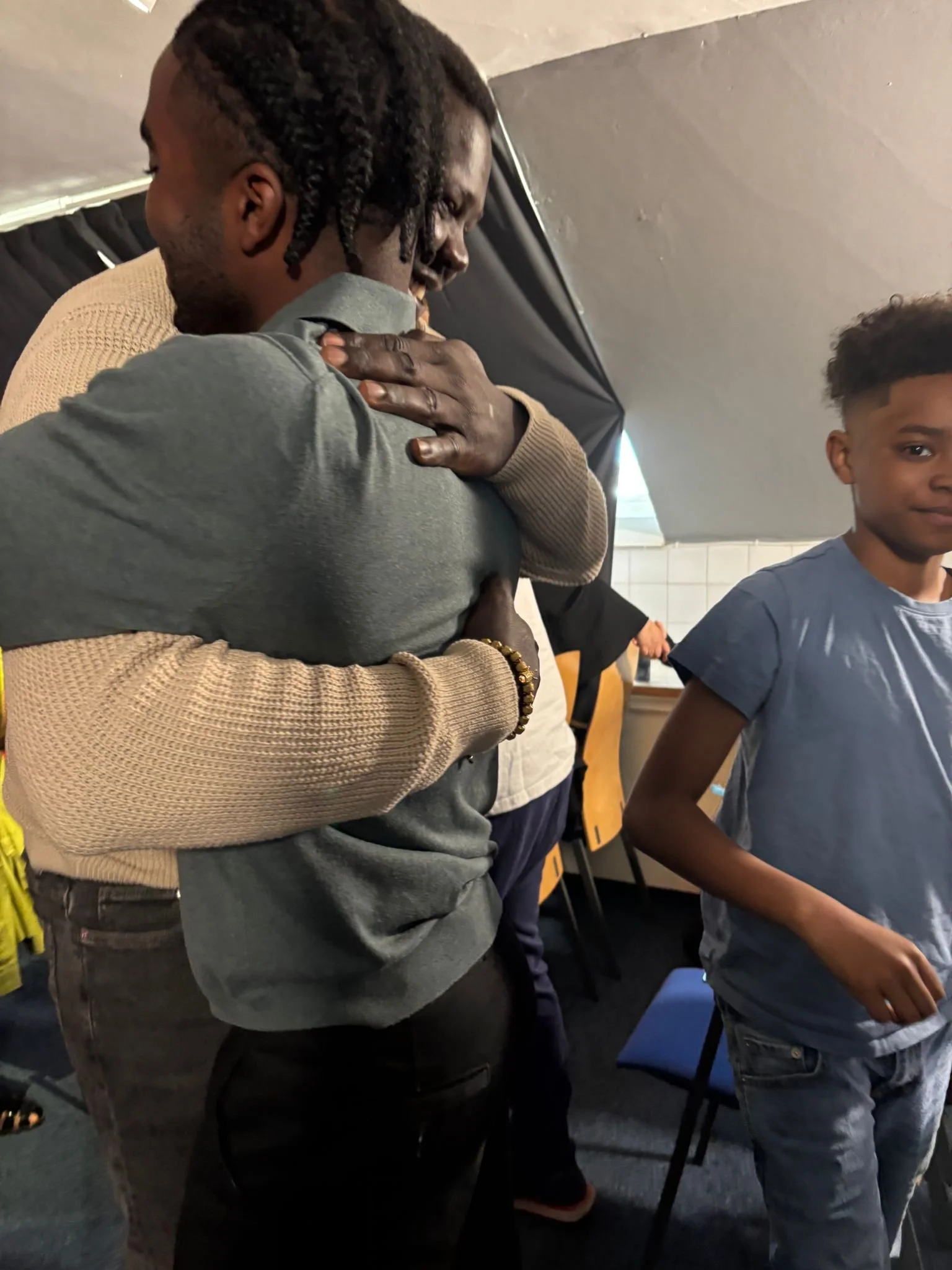 A man with braided hair is hugging a woman in a tan jacket. There are two boys standing nearby, one in the foreground wearing a blue shirt and the other in the background with a yellow shirt. The scene appears to be inside a room with dark walls and 