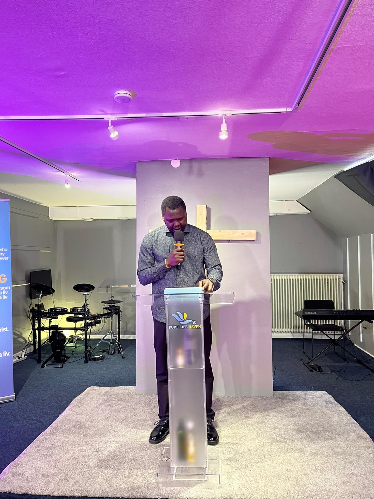 A man standing at a transparent podium with a religious symbol and the words "Pure Life Haven," holding a microphone in a church or community center. There is a cross behind him, musical instruments on his left, and a keyboard on his right. The ceili