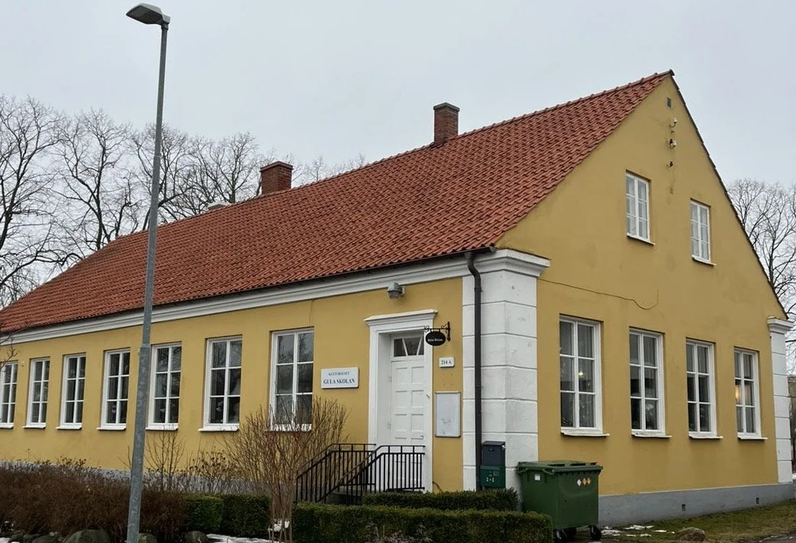 A yellow, two-story building with a red tiled roof and multiple white-framed windows, located on a street with leafless trees in the background, and a streetlamp in the foreground.