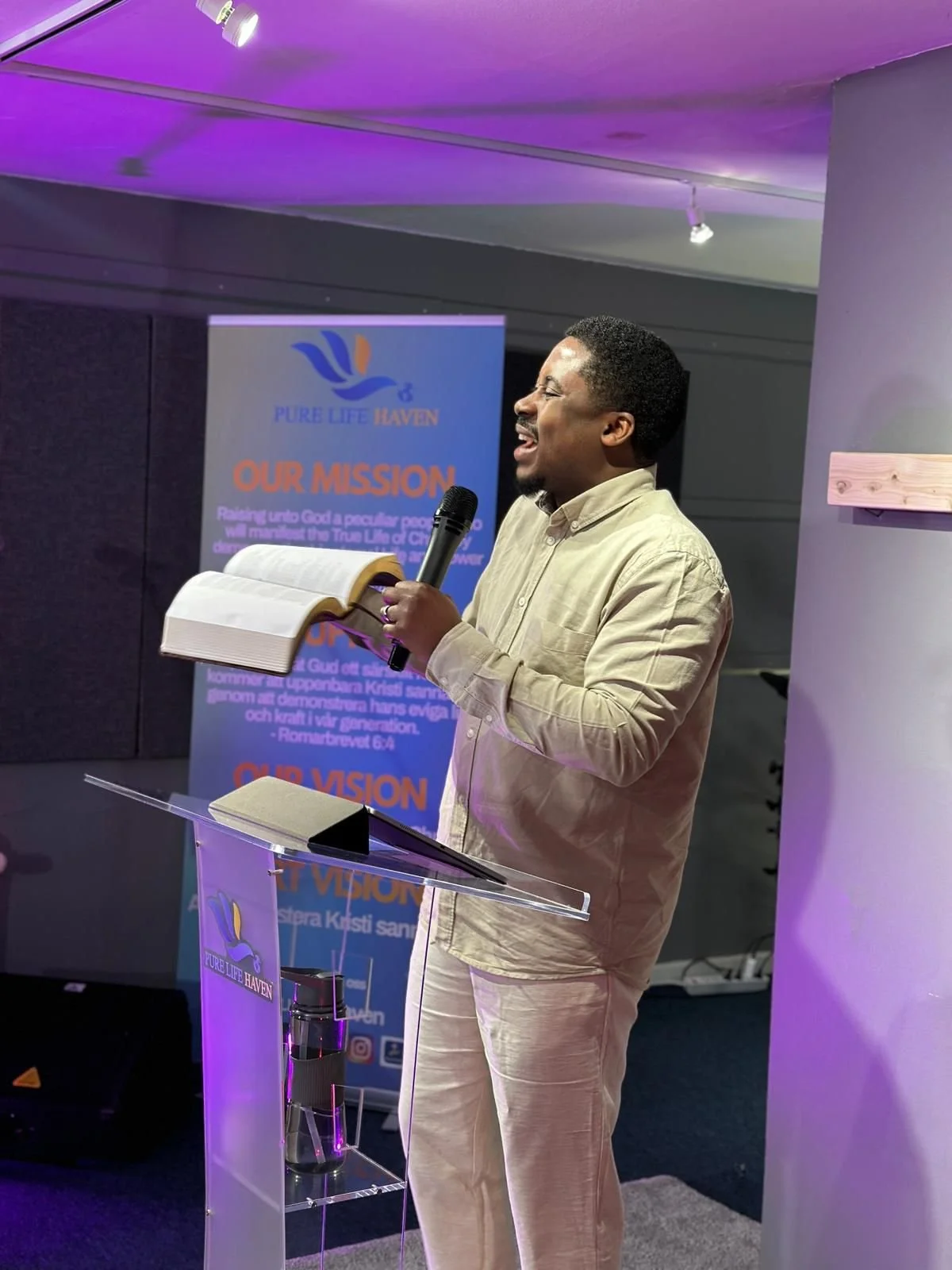 A man holding a microphone and an open book, standing at a podium with a banner behind him that reads 'Our Mission' and 'Pure Life Haven', with purple and blue lighting in the background.