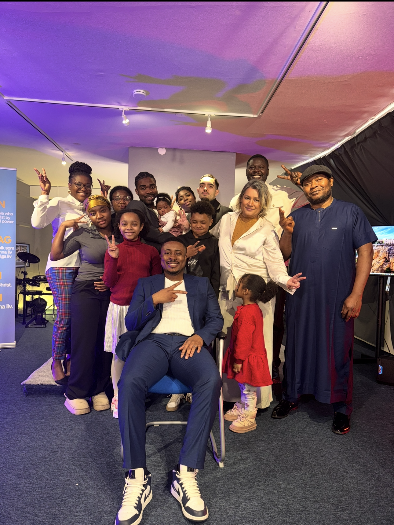 Group photo of diverse people smiling, some holding peace signs, in a room with colorful lighting and a television screen, with children and adults of various ages and backgrounds.