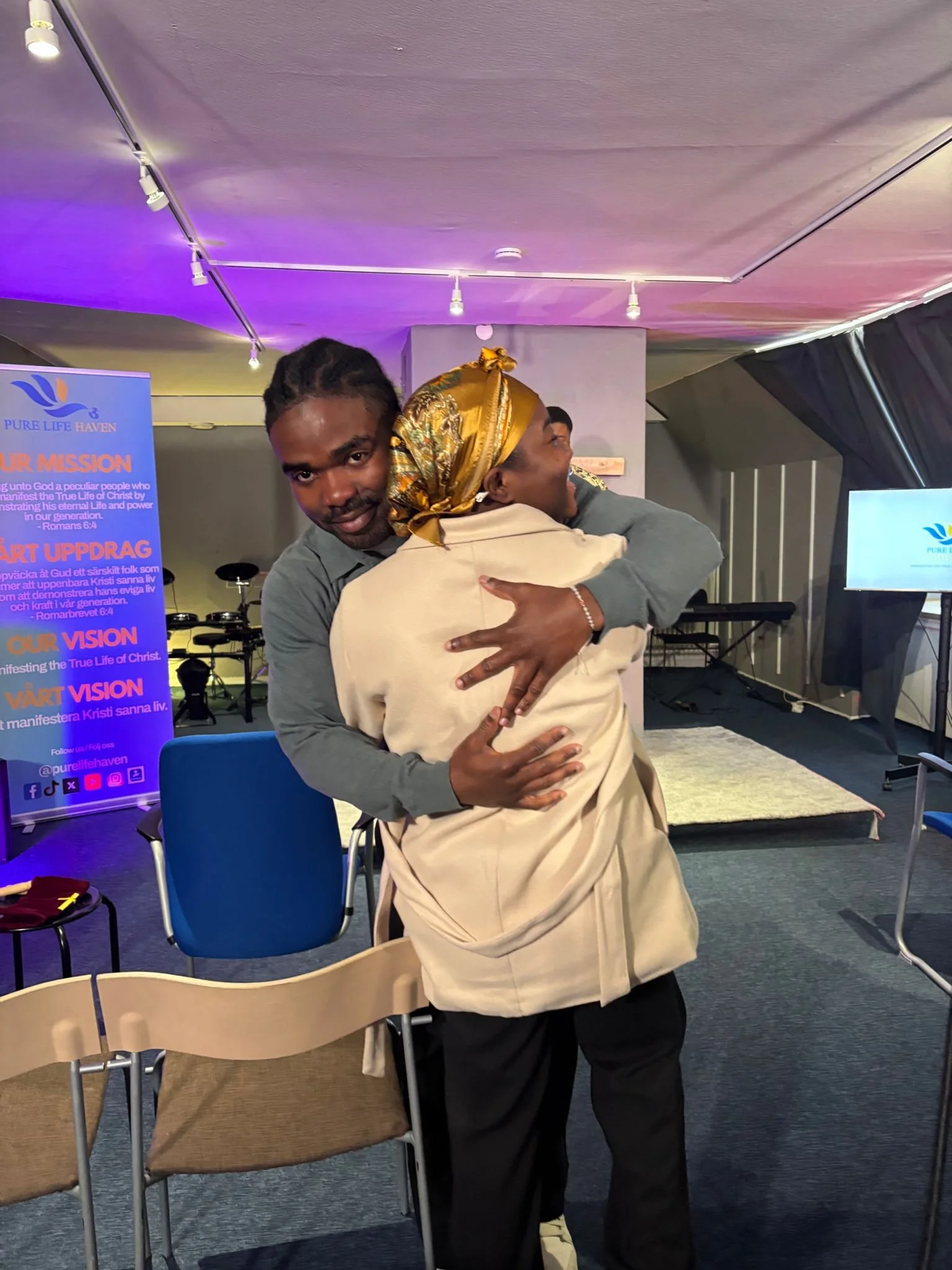 Two people hugging each other at an indoor event, with a man facing the camera and a woman wearing a headscarf. Background includes chairs, a display screen, and a poster for Pure Life Haven.
