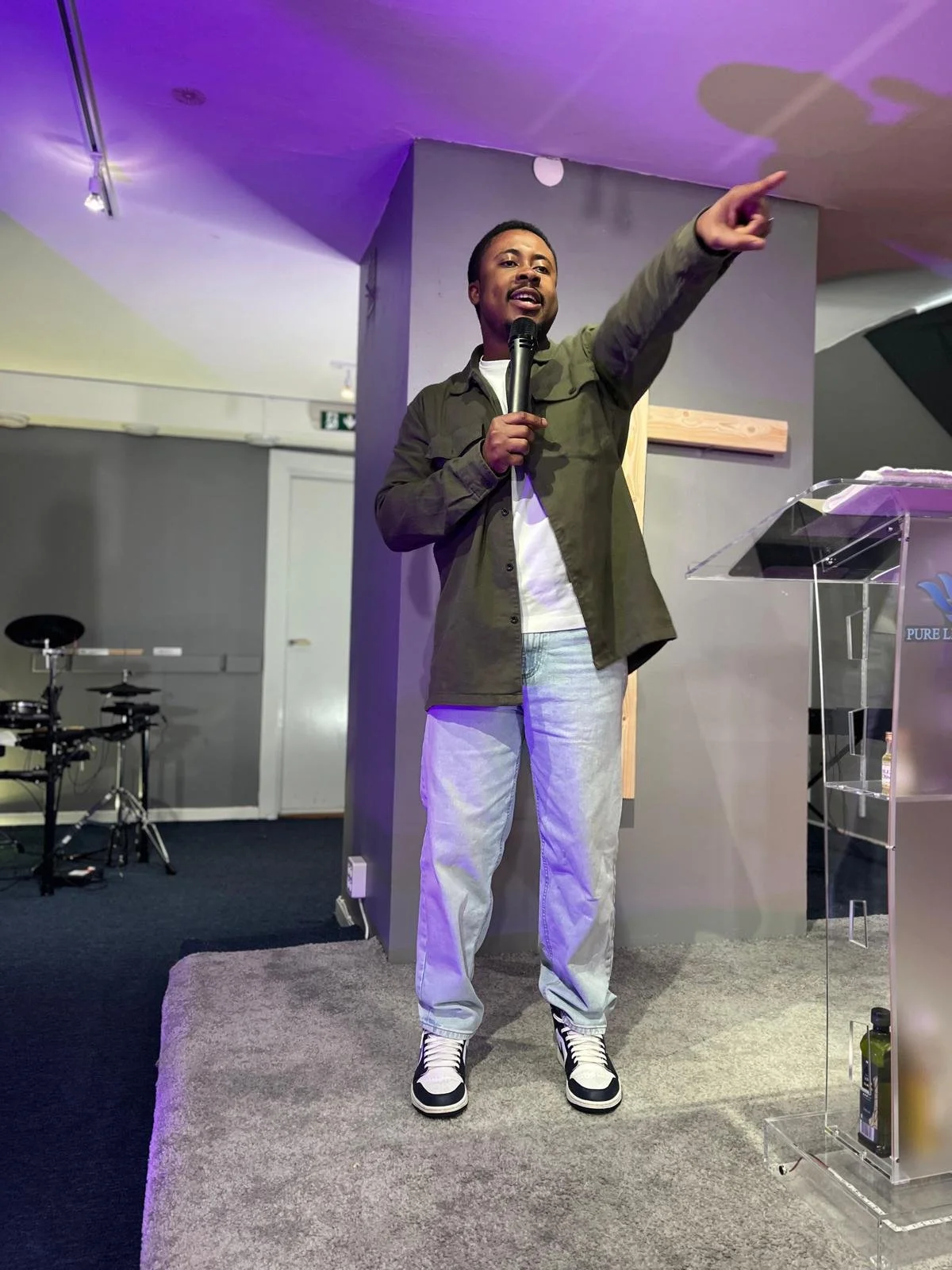 A man standing on a small gray carpeted platform, holding a microphone in his left hand, pointing with his right hand, speaking or presenting in a room with colorful purple and yellow lighting, drums in the background, and a transparent podium to his