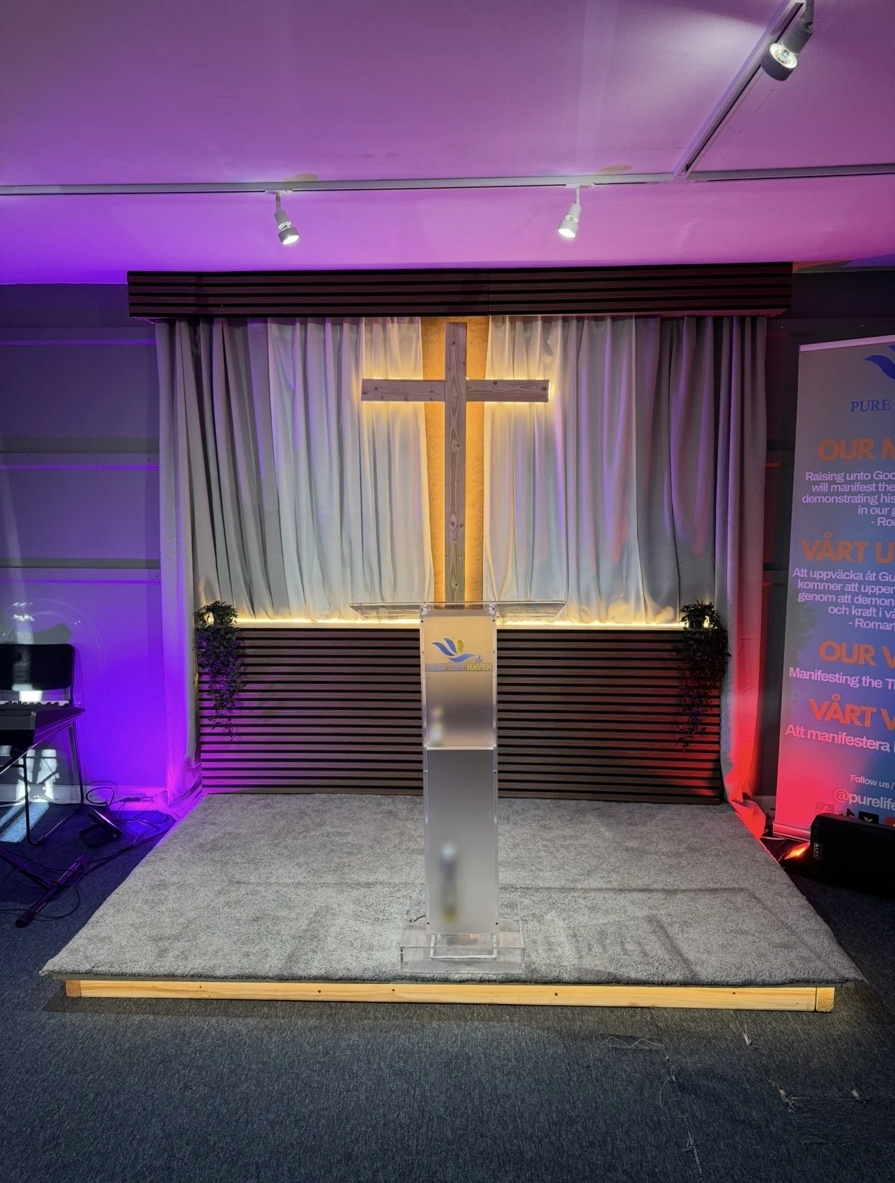 Church stage with illuminated cross and podium, decorated with white curtains, plants on each side, purple and red lighting, and a banner with inspirational text.