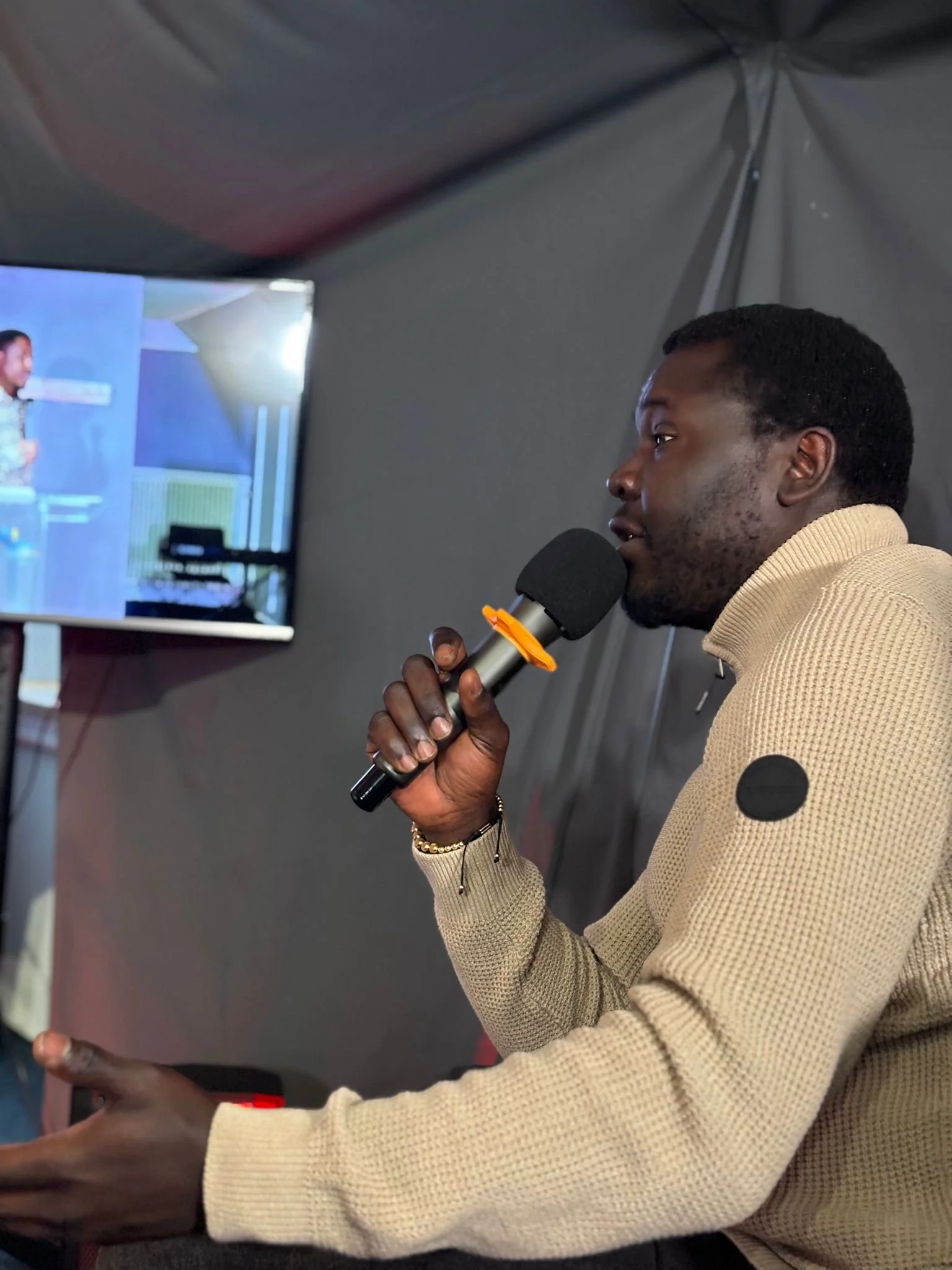 A man speaking into a microphone while looking at a screen displaying another person, inside a dark room or tent.