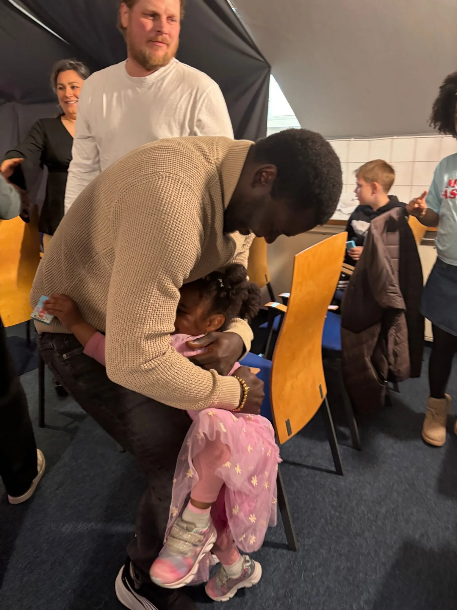 A man in a tan checkered jacket hugs a young girl in pink pajamas with star designs, while standing next to a row of chairs in a room with several other people, including a woman and a boy holding a phone.