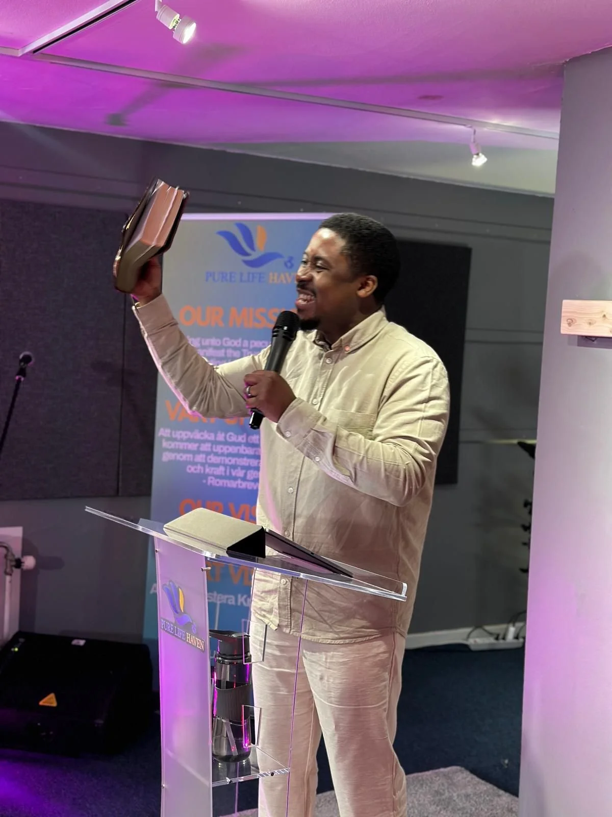A man wearing a beige shirt and light-colored pants is speaking into a microphone while holding a book in the air. He is standing at a transparent lectern with a logo that says 'Pure Life Haven' and a lit-up background with a banner matching the same