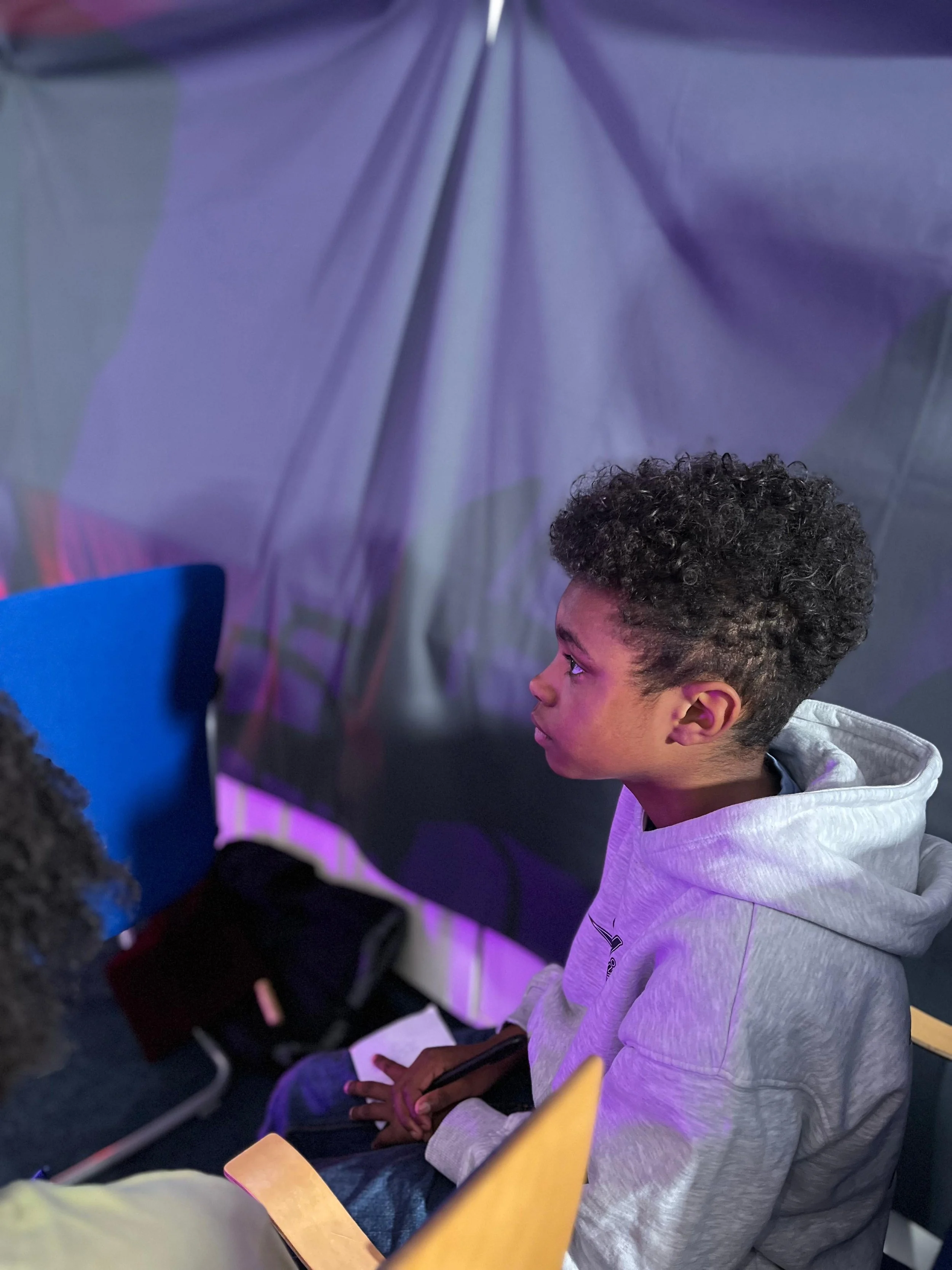 A young person with curly hair is sitting indoors, wearing a light gray hoodie, with a thoughtful expression, surrounded by purple and blue lighting.