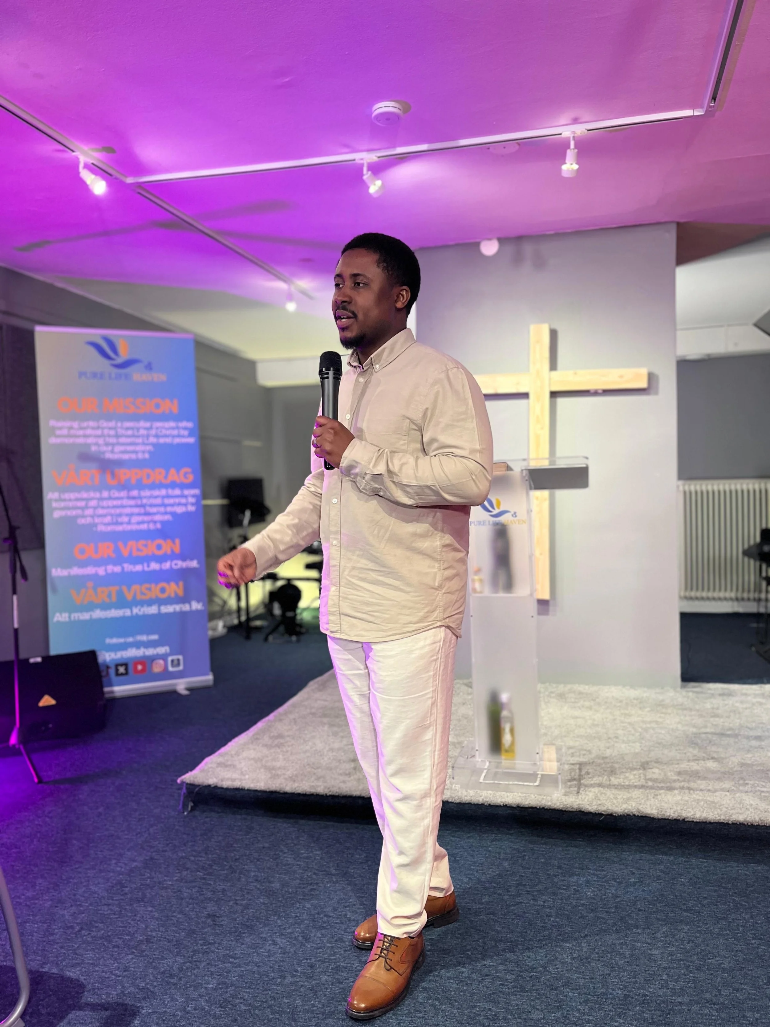 A man with short black hair and a mustache, dressed in a beige button-up shirt and white pants, is speaking into a microphone in what appears to be a church or conference setting. Behind him is a large wooden cross on the wall, and a purple-lit ceili