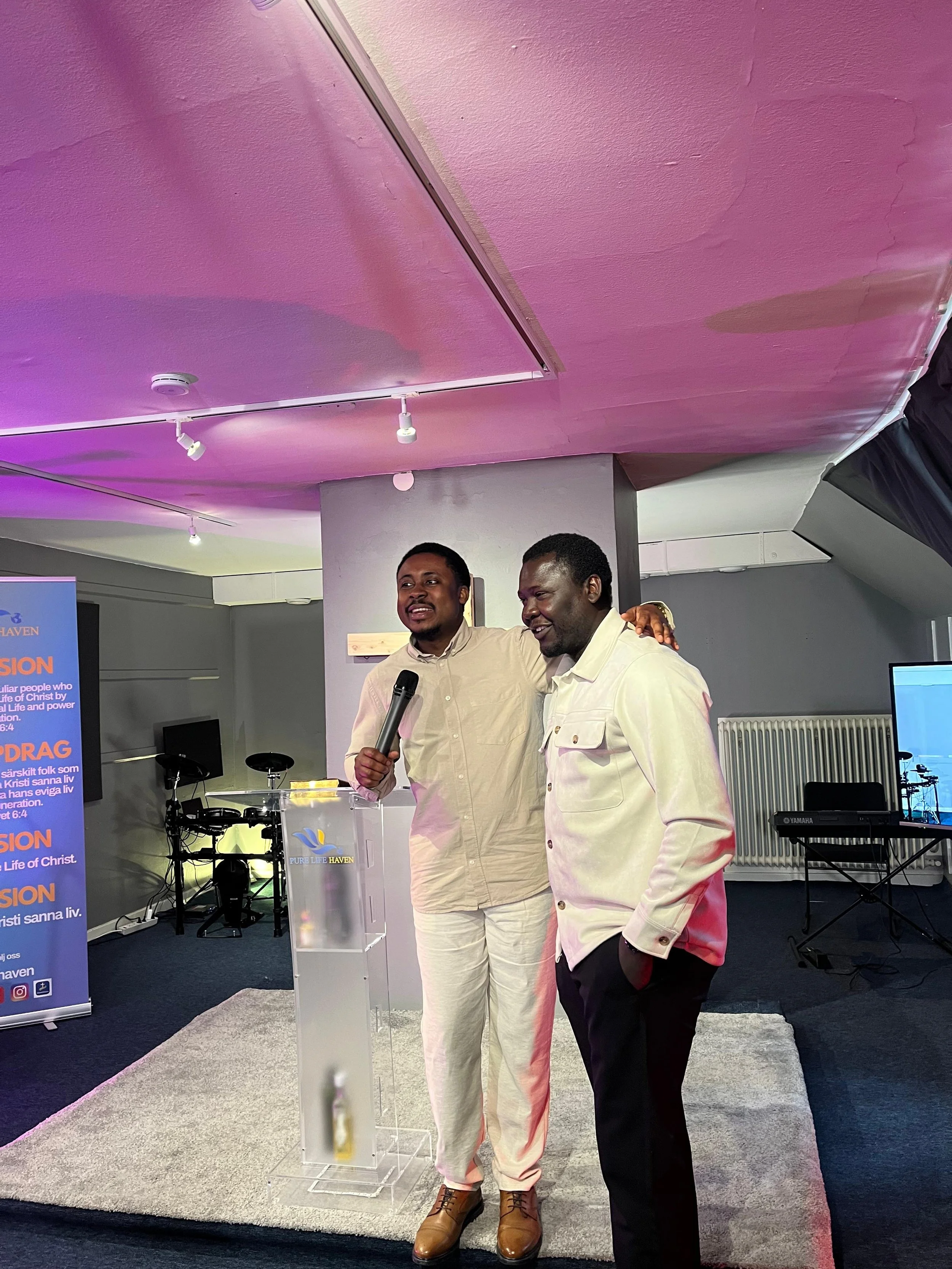 Two men standing on a stage or platform, smiling and posing for a photo. One is holding a microphone. Background includes a Bible verse poster, electronic equipment, and a podium with a logo that says 'Pure Life Haven.' The setting appears to be a ch