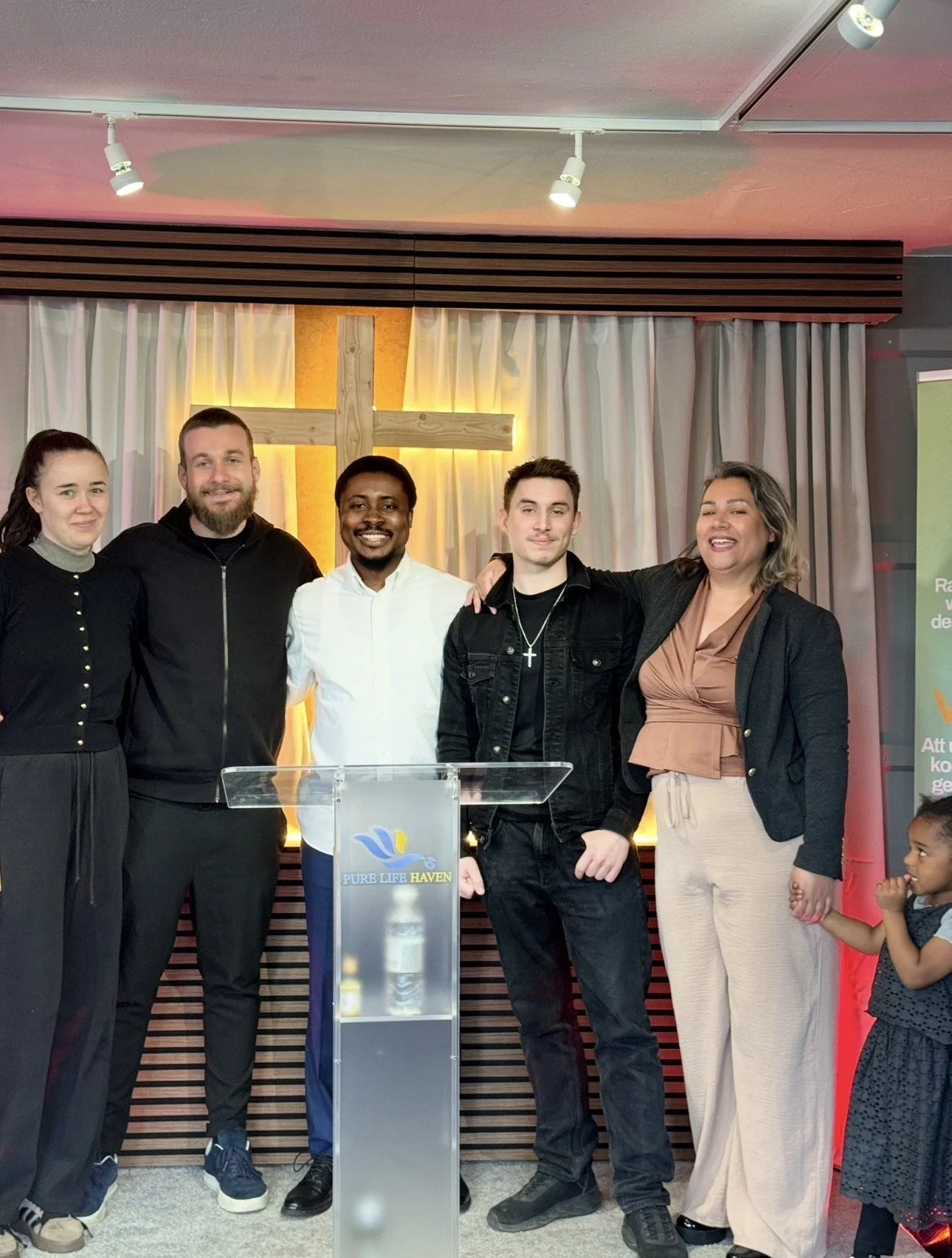 A diverse group of five people standing together behind a clear podium with a church cross in the background, inside a church or religious setting.
