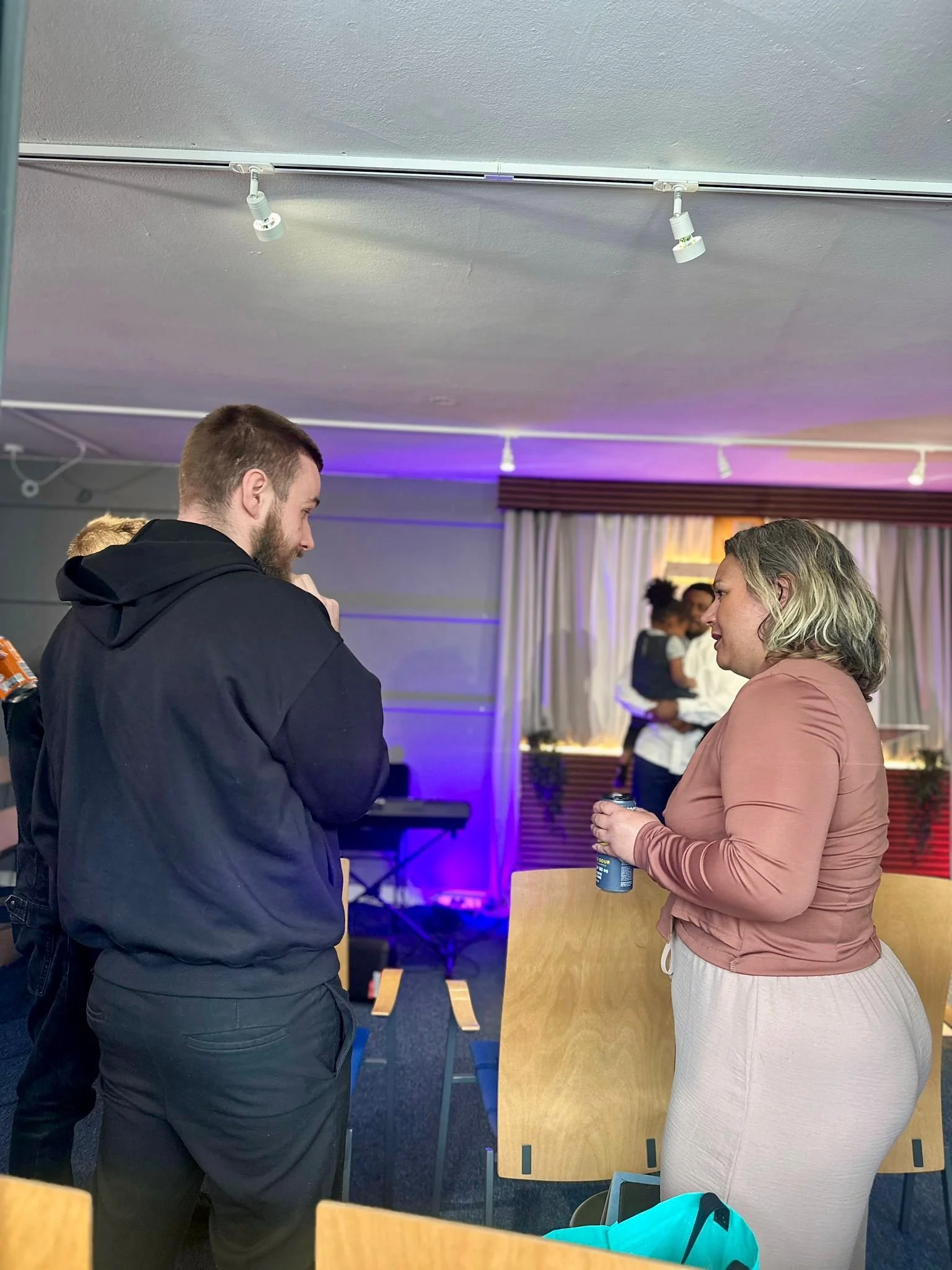 Two people, a man and a woman, are standing and talking in a room with a few tables and chairs, some people are blurred in the background, and there are curtains and colored lighting on the wall.