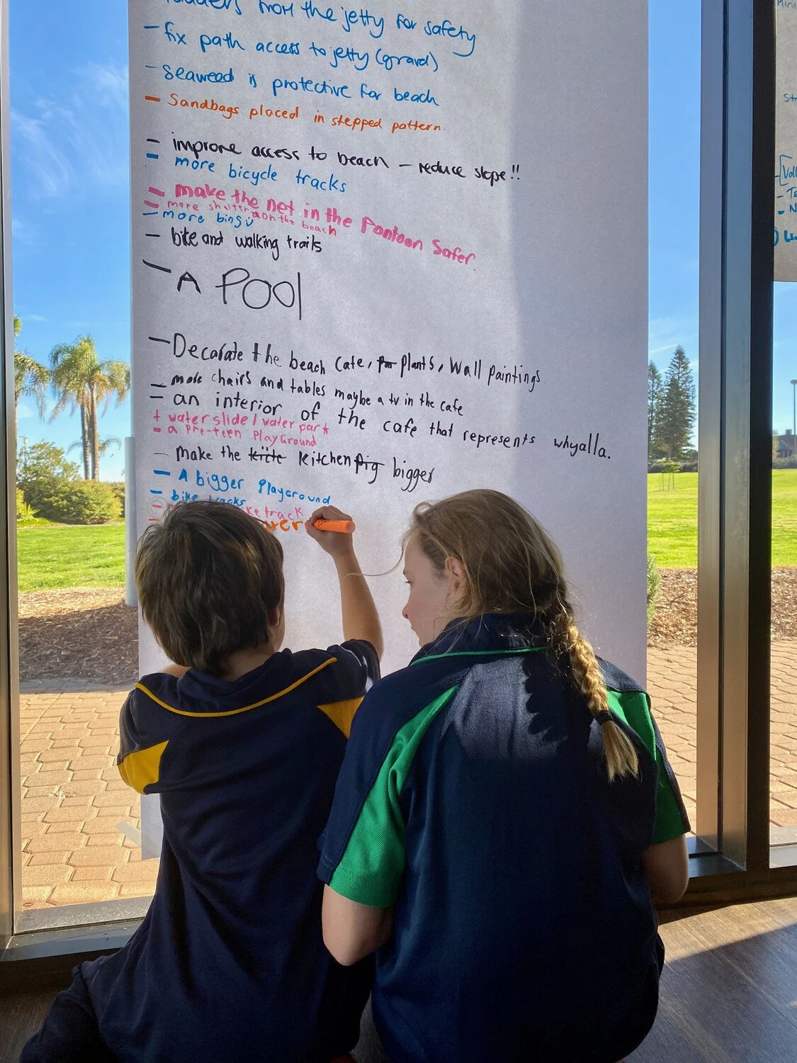 Two children, a boy and a girl, are writing on a large white poster board with a black marker. The poster contains handwritten notes about beach safety and a pool. They are inside a building near a window with a view of trees, grass, and a sunny sky outside.