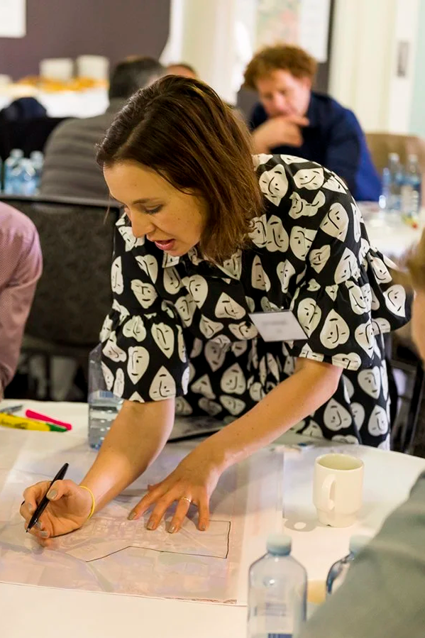 Liesl Codrington as lead consultant with Vaere Social. She has shoulder-length brown hair wearing a black and white patterned dress is leaning over a table, writing or drawing on a large sheet of paper.