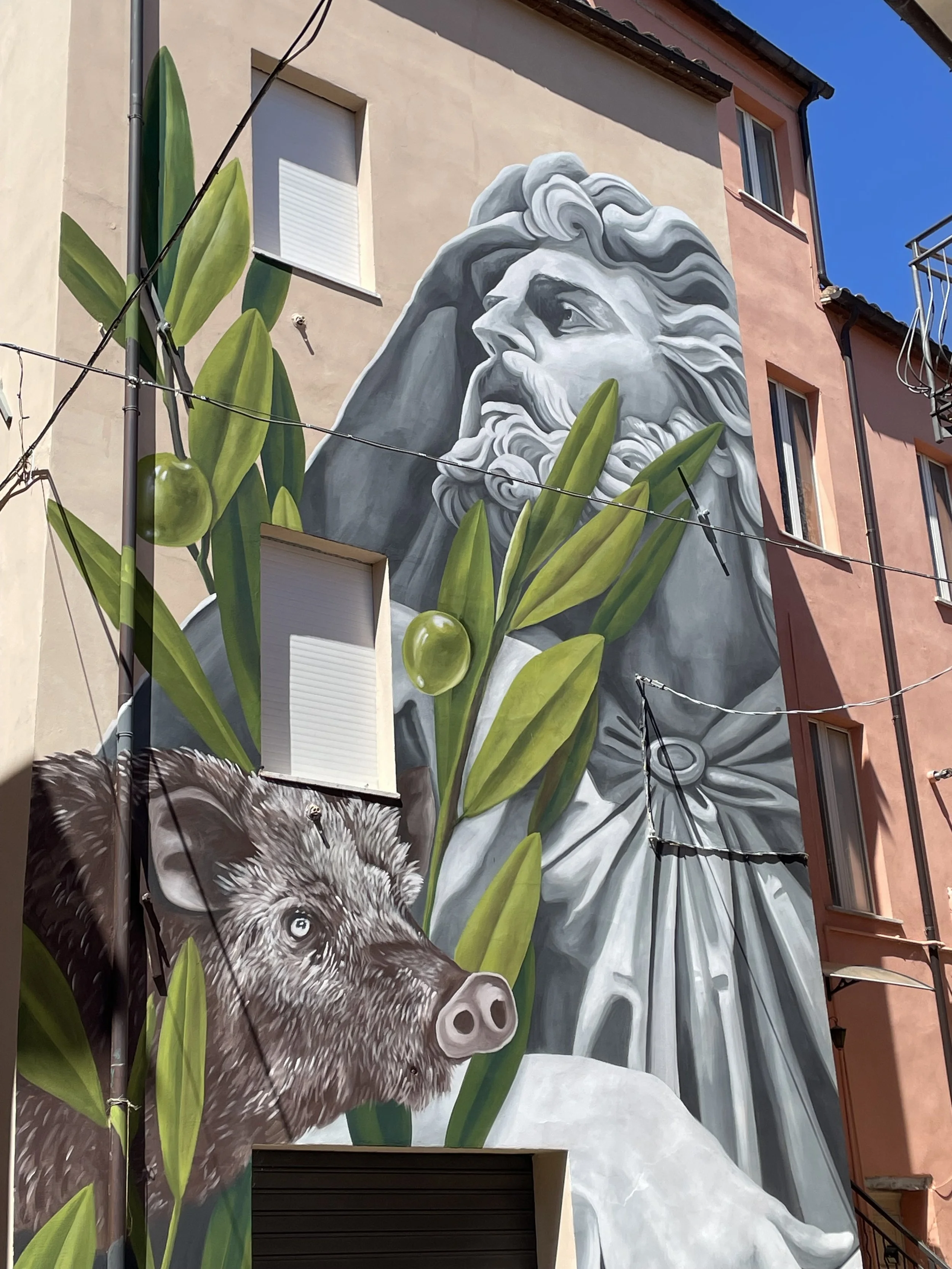 Mural featuring a grayscale portrait of a bearded ancient Greek or Roman figure, with a boar and green olive branches in the foreground, painted on the side of a building.