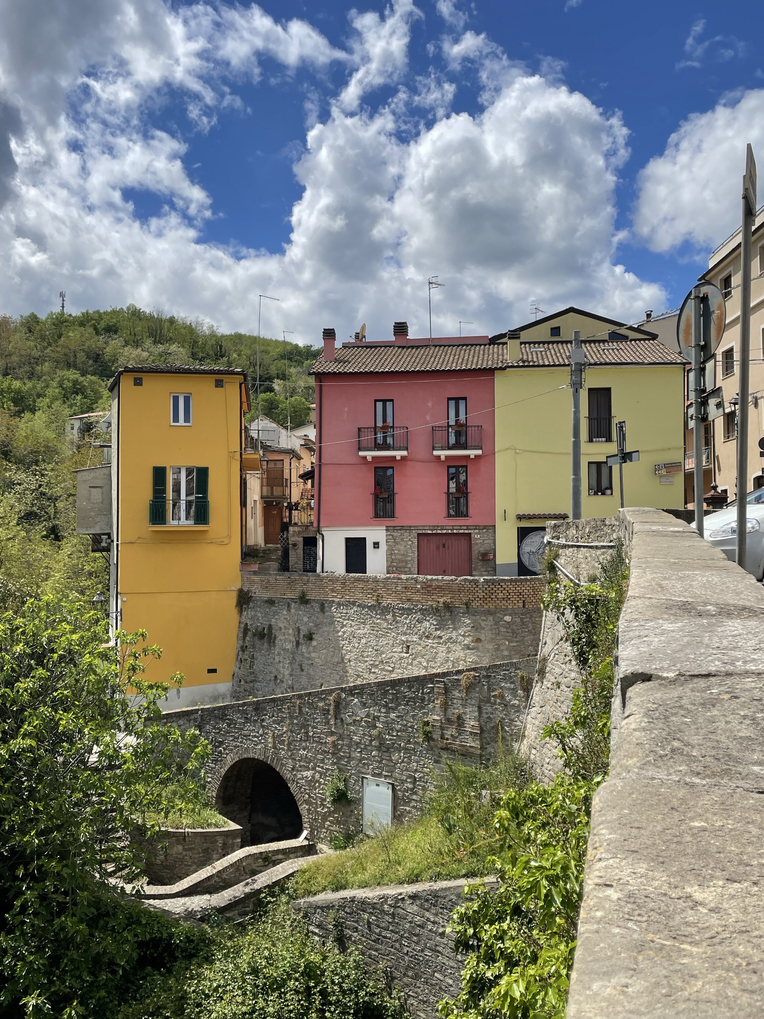 Colourful buildings on a hillside with a stone bridge and lush greenery under a partly cloudy sky.