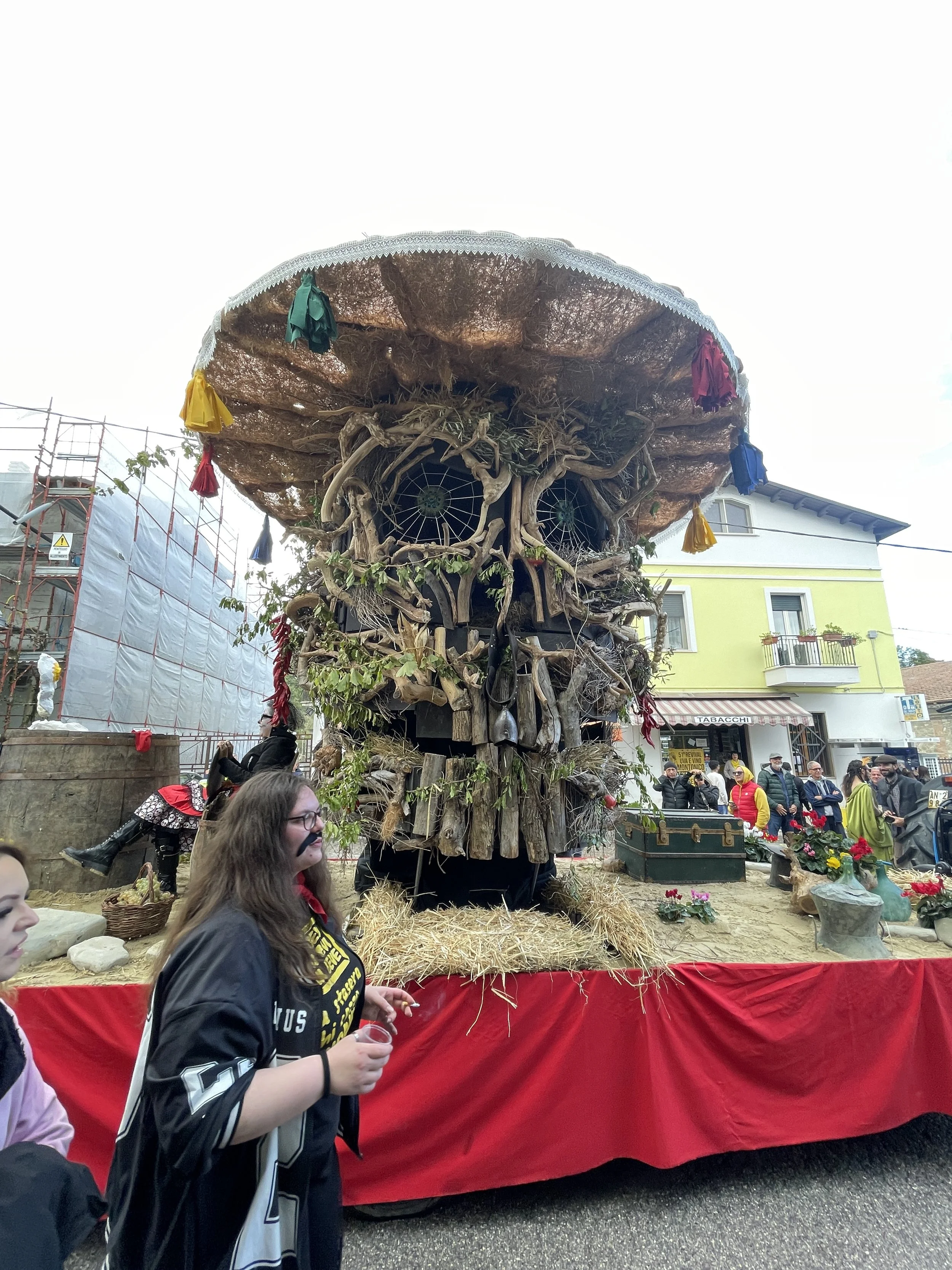 Large wooden sculpture of a face with eyes, nose, and mouth made from twisted wood and branches, decorated with colorful cloths, displayed on a platform with straw, in a street festival setting.