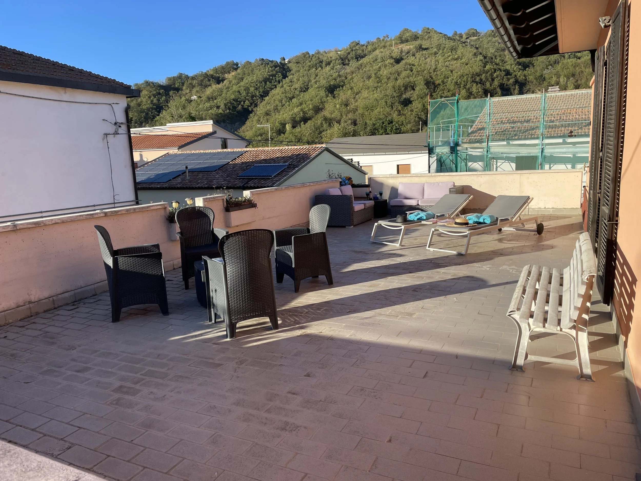 Outdoor terrace with black wicker chairs around a table, sun loungers with blue towels, pink cushioned outdoor sofa, wooden bench, and potted plants, overlooking rooftops and green hills under a clear blue sky.