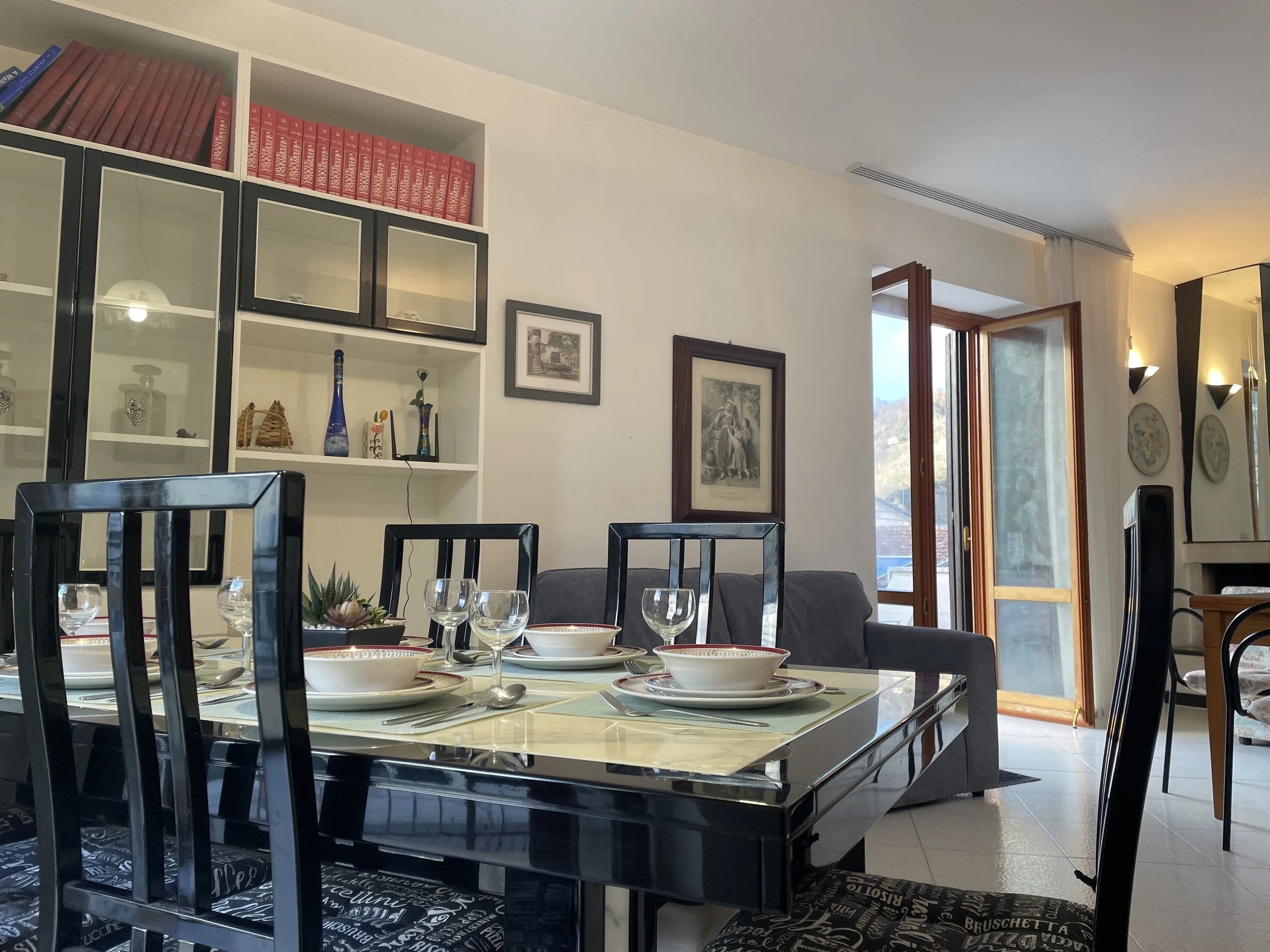 Dining room with table set for meal, black chairs, white shelves with books and decorations, open door to balcony, and wall decorations.