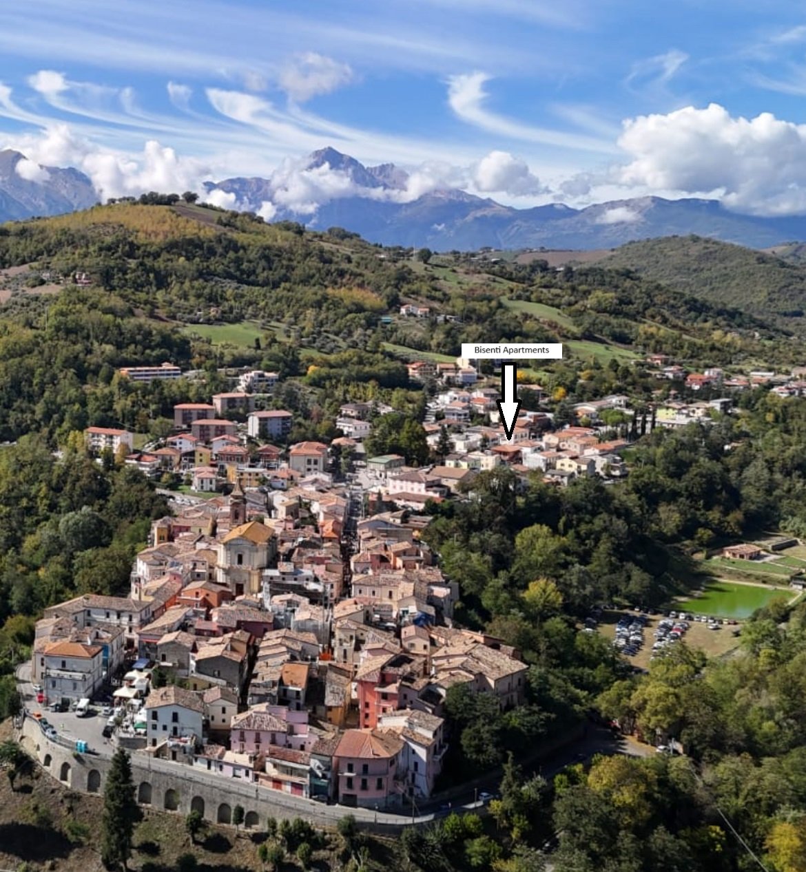A scenic view of a hillside town with colorful buildings, surrounded by lush green trees, with distant mountains and a partly cloudy sky in the background. An arrow labels the Bisenti Apartments.