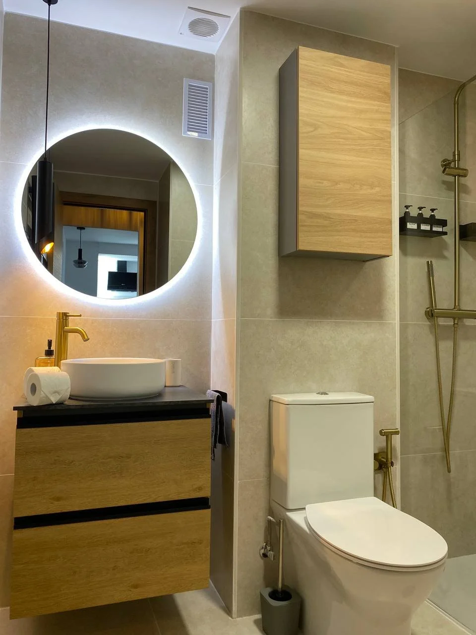 Modern bathroom with wall-mounted wooden vanity with vessel sink, round mirror with backlit LED, toilet, wooden wall cabinet, and shower with gold fixtures.