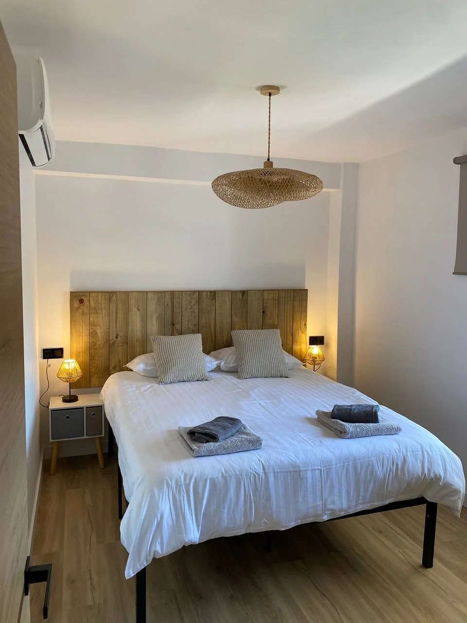 A neatly made bed with white linens, two striped pillows, and two folded towels on top. Two bedside lamps with warm lighting are on either side of the bed, and a wooden headboard extends across the width. The room has light-colored walls, a wooden fl