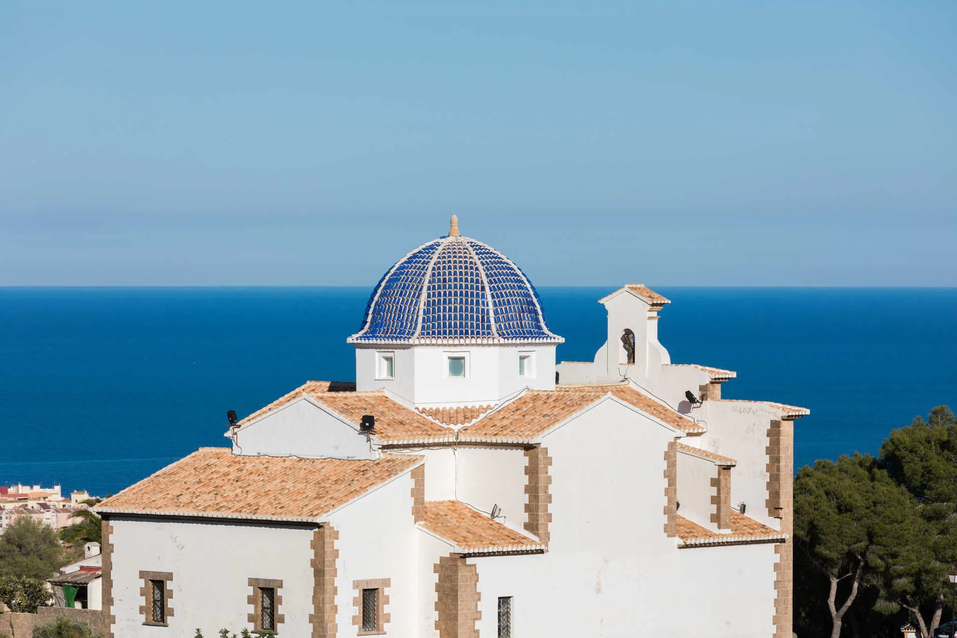 White church with blue tiled dome roof overlooking the ocean and surrounded by trees.
