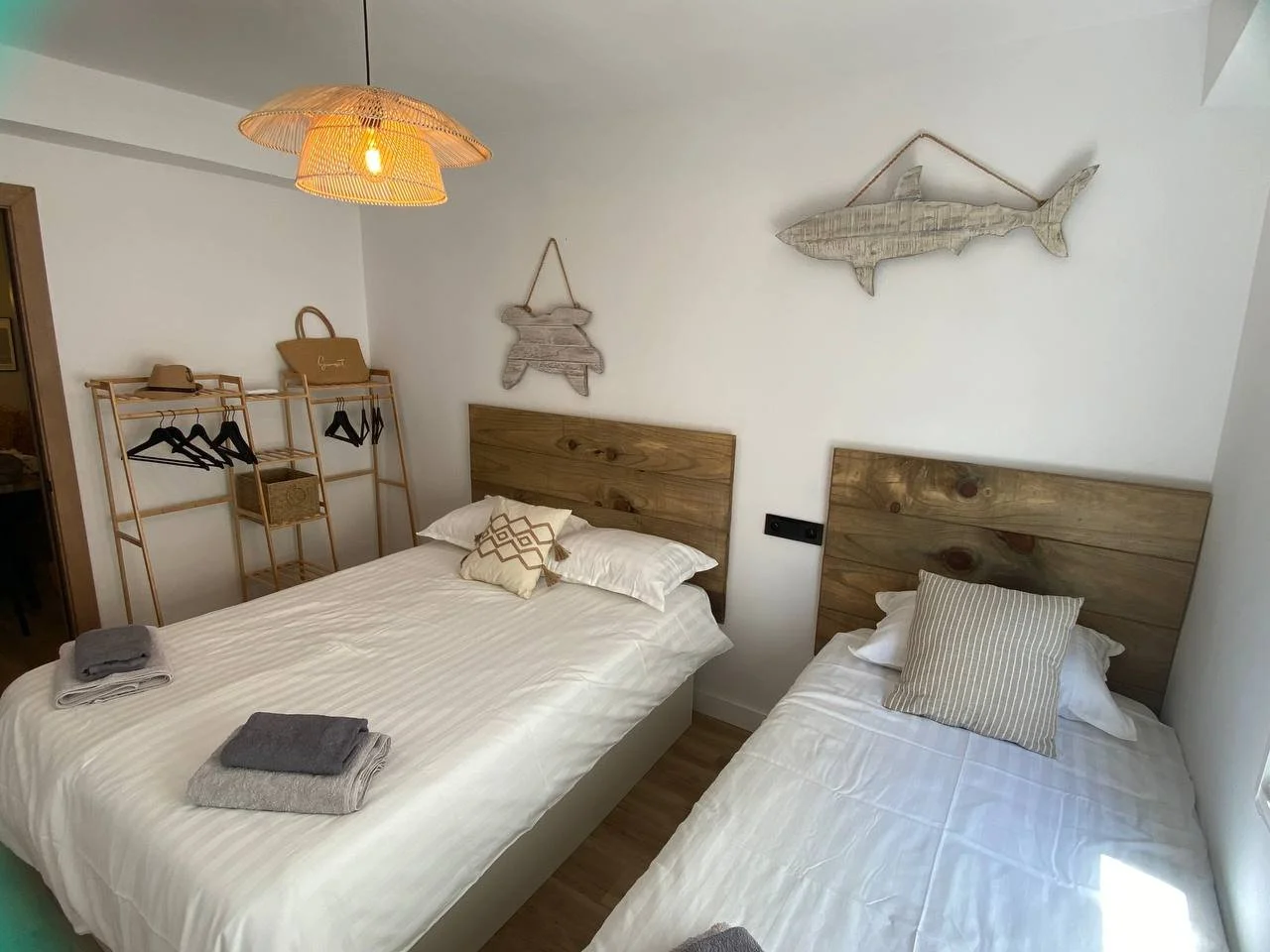 Hotel bedroom with two beds featuring wooden headboards, a hanging fish and fish-shaped wall decor, a rattan light fixture, and a wooden clothing rack with hats and hangers.