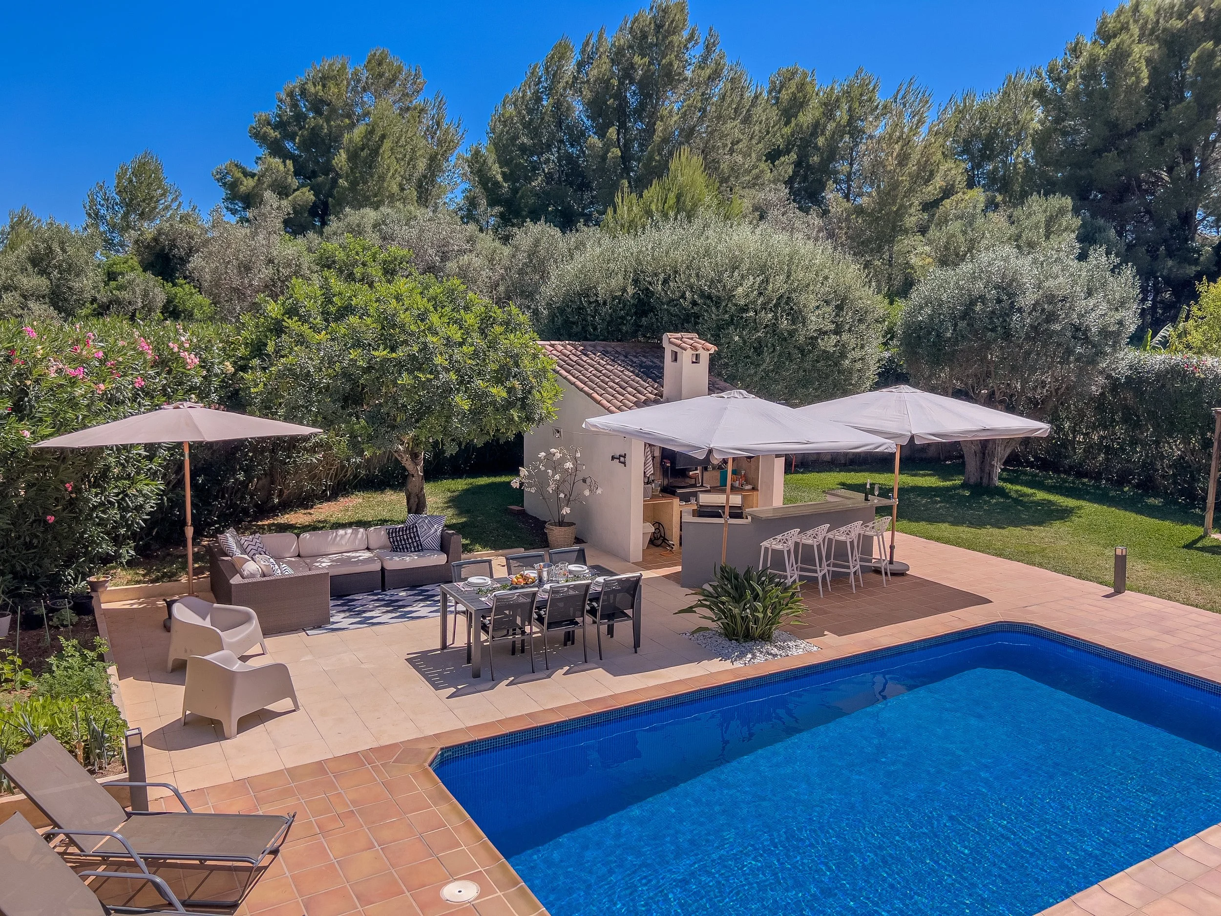 Backyard with swimming pool, outdoor seating, umbrellas, and lush greenery under a clear blue sky.