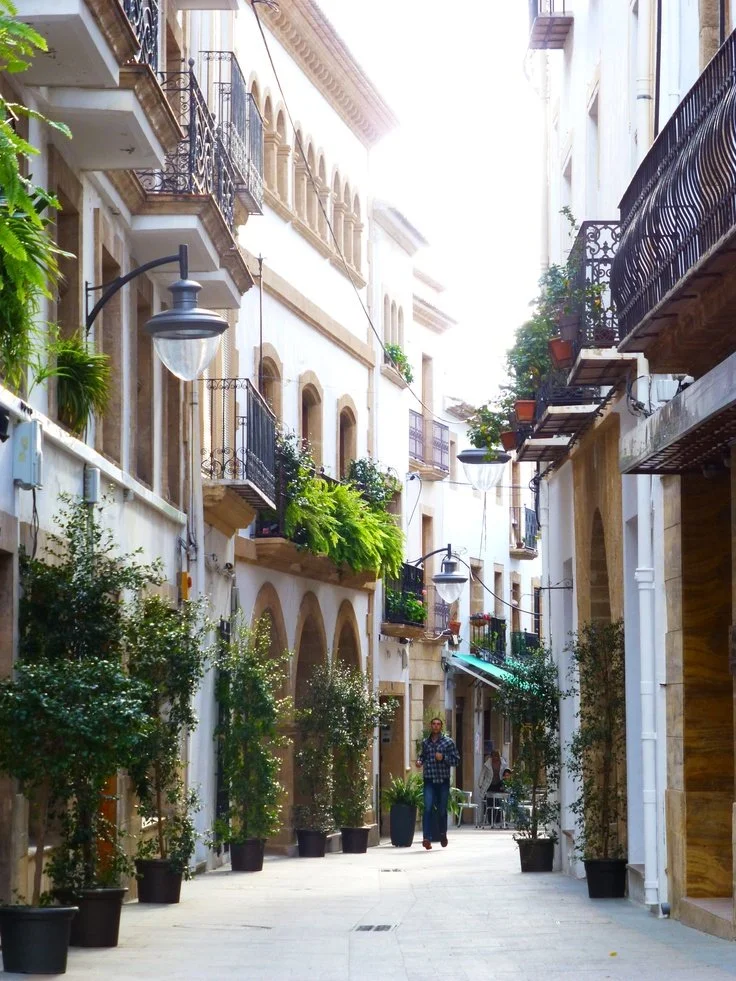 A narrow street lined with multi-story buildings with balconies, potted plants, and street lamps. A person is walking in the street, which has potted plants and outdoor seating.