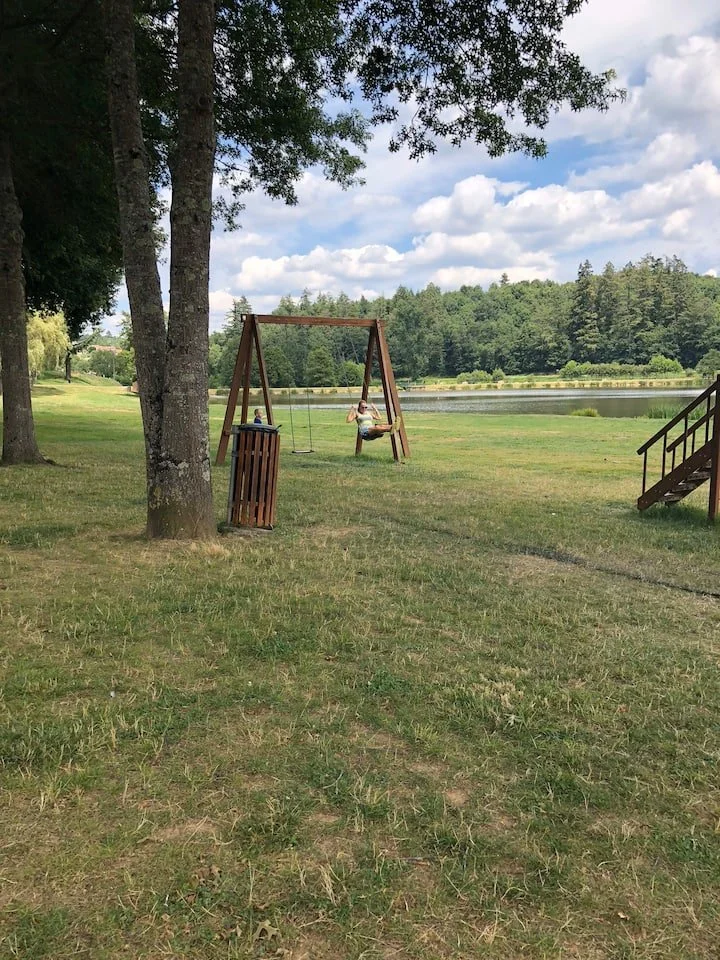 A grassy park with a lake in the background, a swing set with one child sitting on the swing, a trash can, and wooden stairs on the right side.
