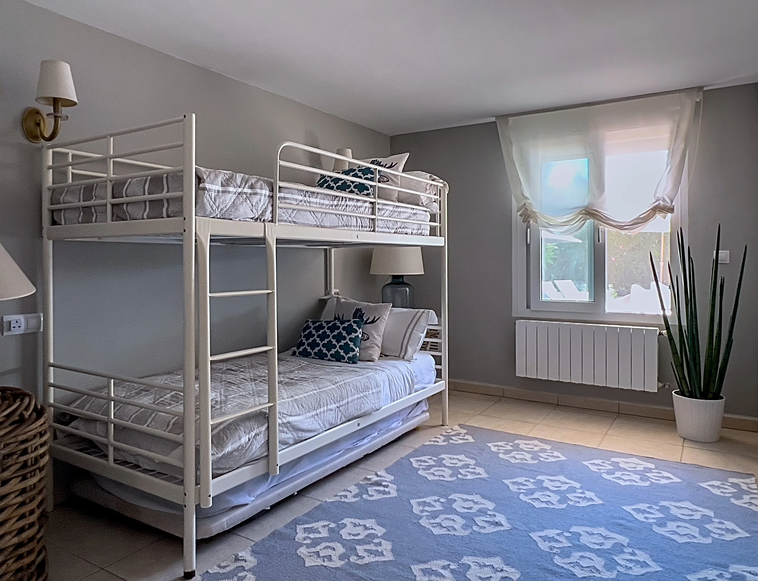A bedroom with a white bunk bed against a light gray wall, two patterned blue and white pillows, a window with sheer white curtains, a large potted plant, a blue and white patterned rug, and a wicker basket.