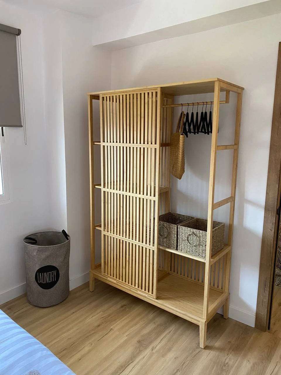 Wooden open wardrobe with hanging bar, two woven baskets, and a tote bag inside; laundry basket labeled "LAUNDRY" in the corner; room with wooden flooring and a window with a roller shade.